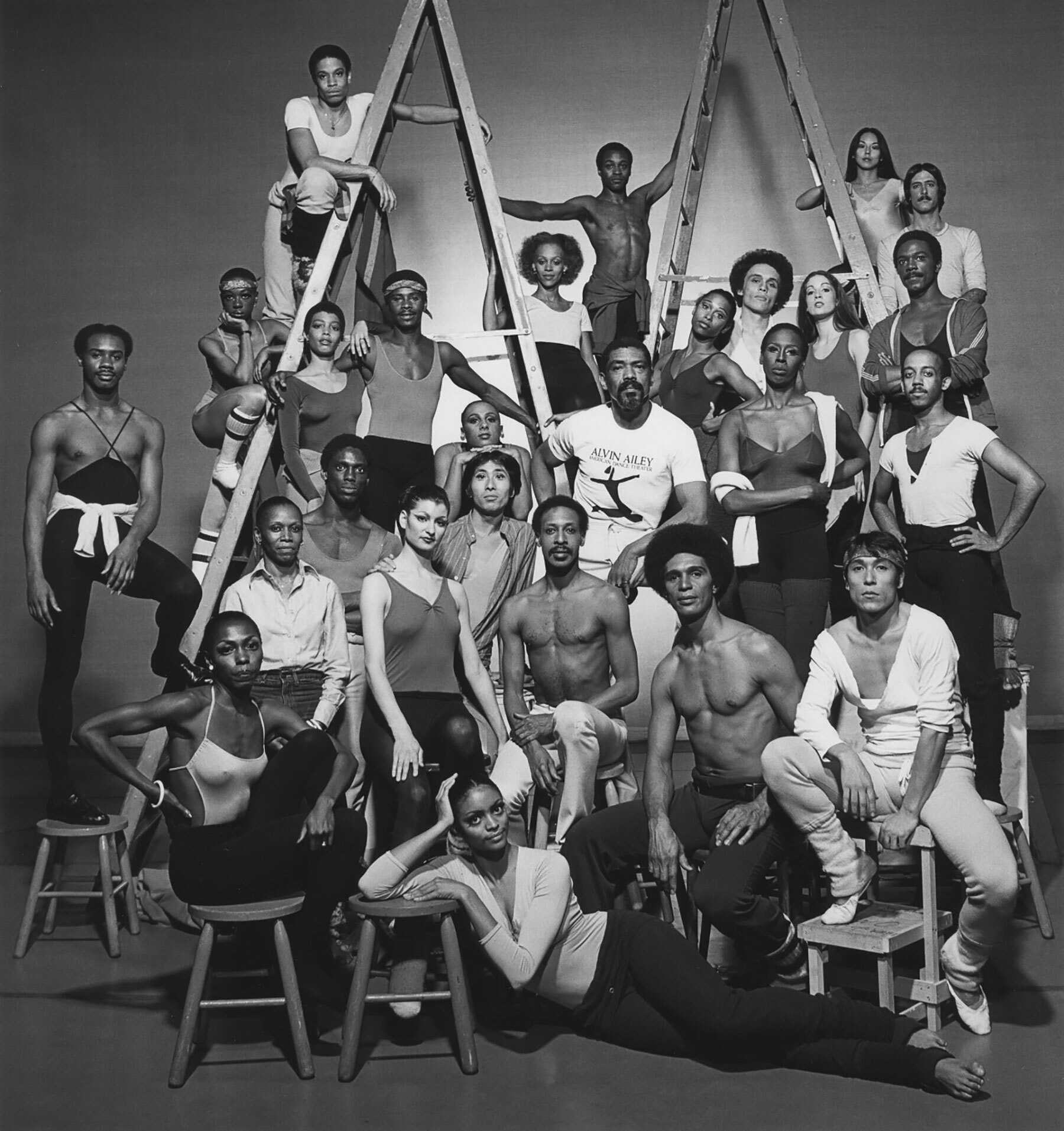 Group portrait of dancers in front of a lattice of ladders, some seated and others standing.