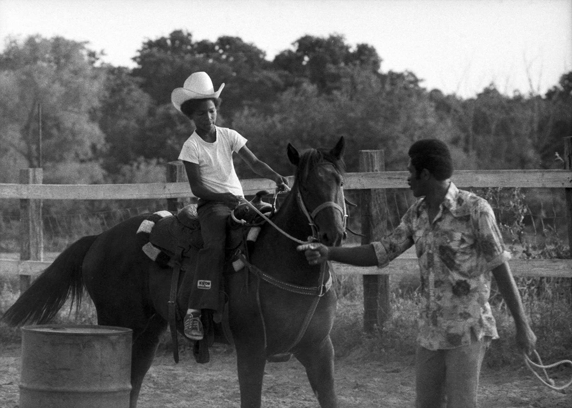 Black-and-white photo of a child wearing a cowboy hat, riding a horse, with an adult guiding the horse in a fenced area.