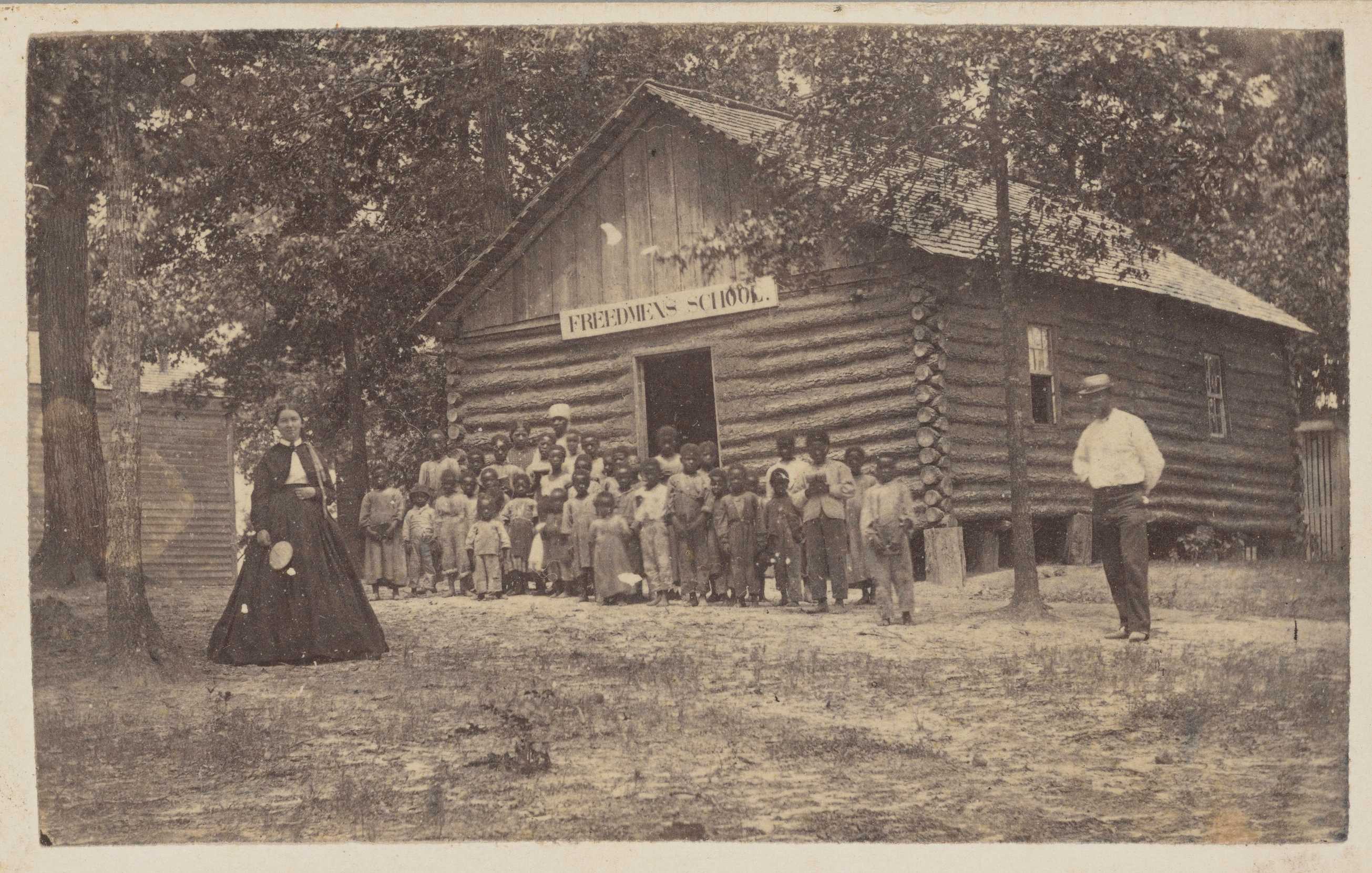 A black and white photo of a teacher and her students outside of a logged cabin building that is their school.