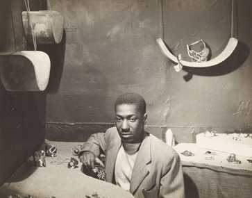 Black-and-white photo of a man seated at a table displaying various jewelry pieces in a small workshop setting.