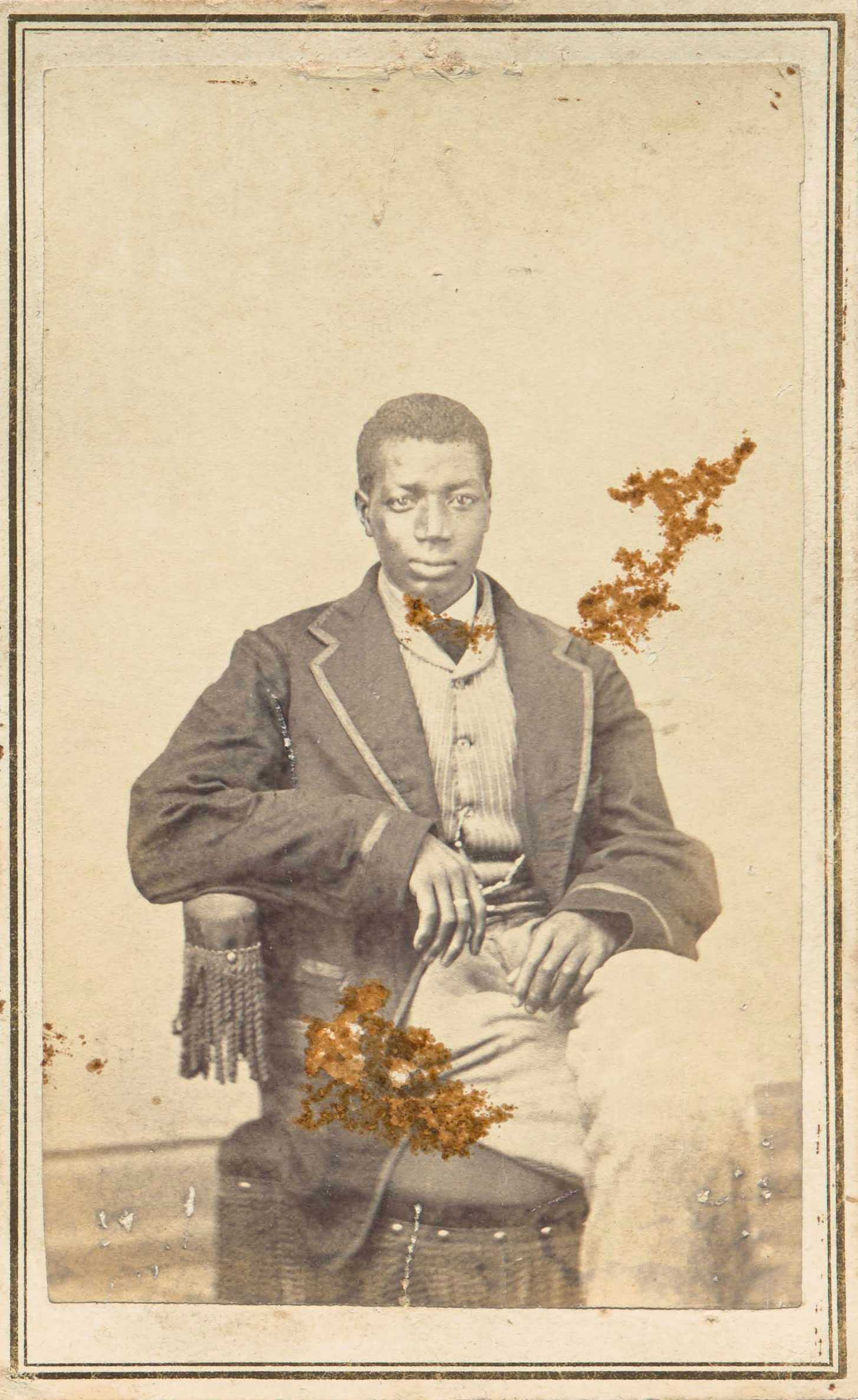 Sepia-toned photograph of a seated young man in formal attire, looking directly at the camera, with light marks on the image.
