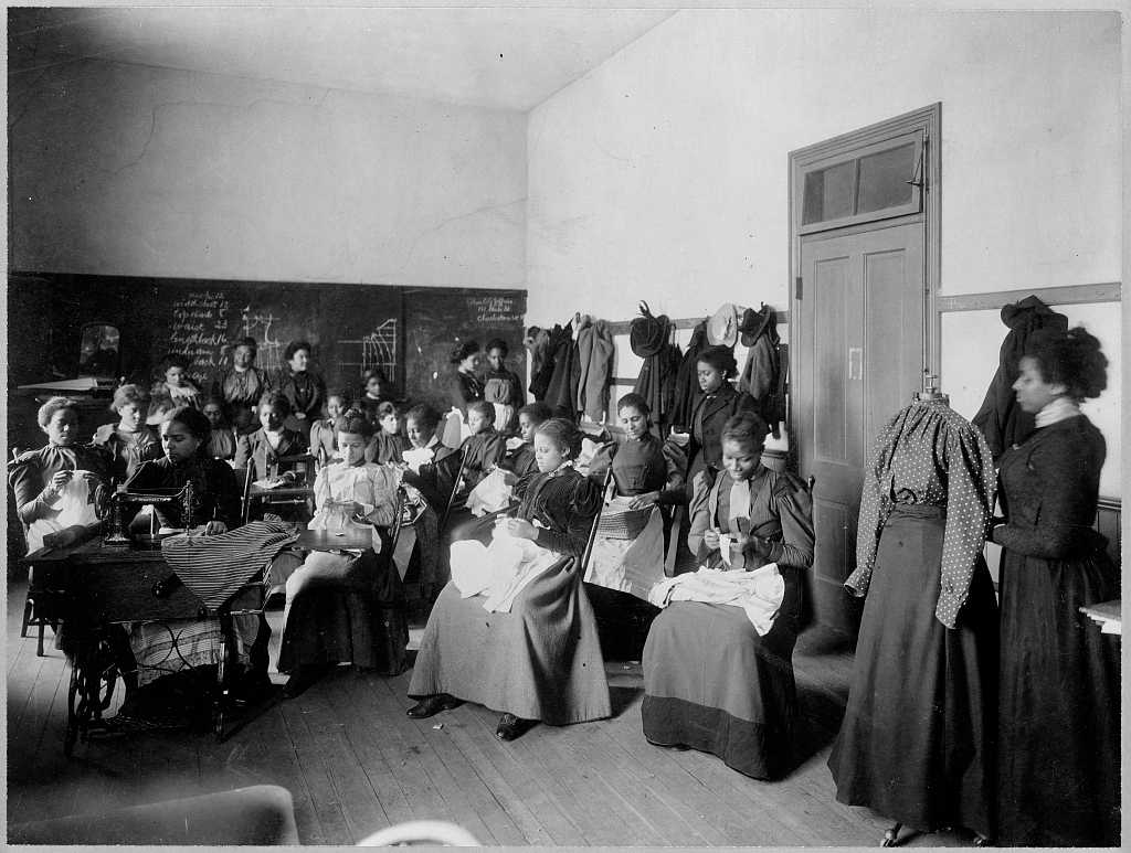 Black and White photograph of female African American students seated in a classroom.  They appear to be sewing/  There are several women standing toward the chalkboard in the rear of the class.  There is one sewing machine in the foreground, being used by a woman.  A woman stands in the front of the class next to a dressed mannequin wearing a long-sleeved blouse and long skirt.