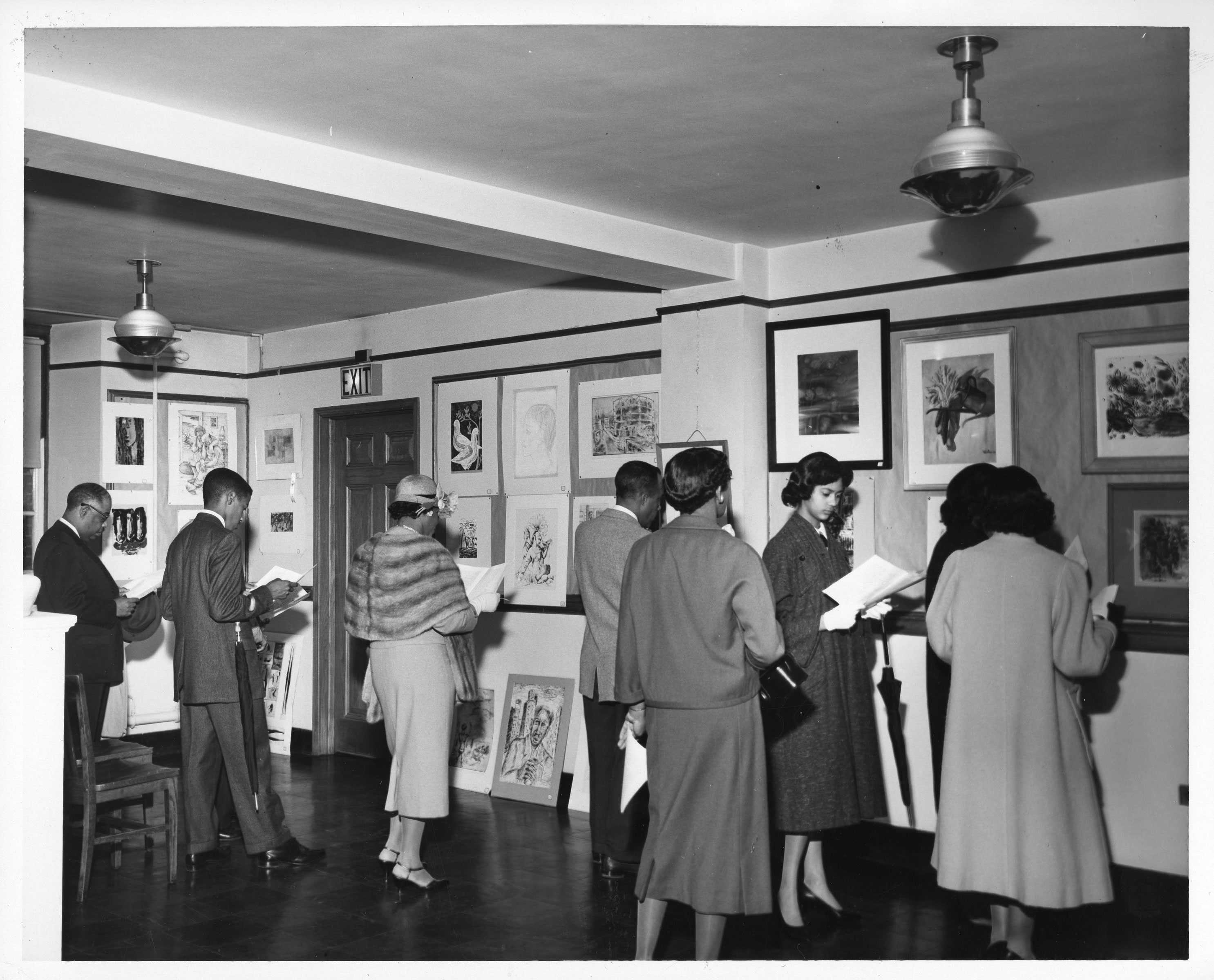 Black-and-white photo of people viewing art in a gallery setting, with various framed artworks displayed on the walls.