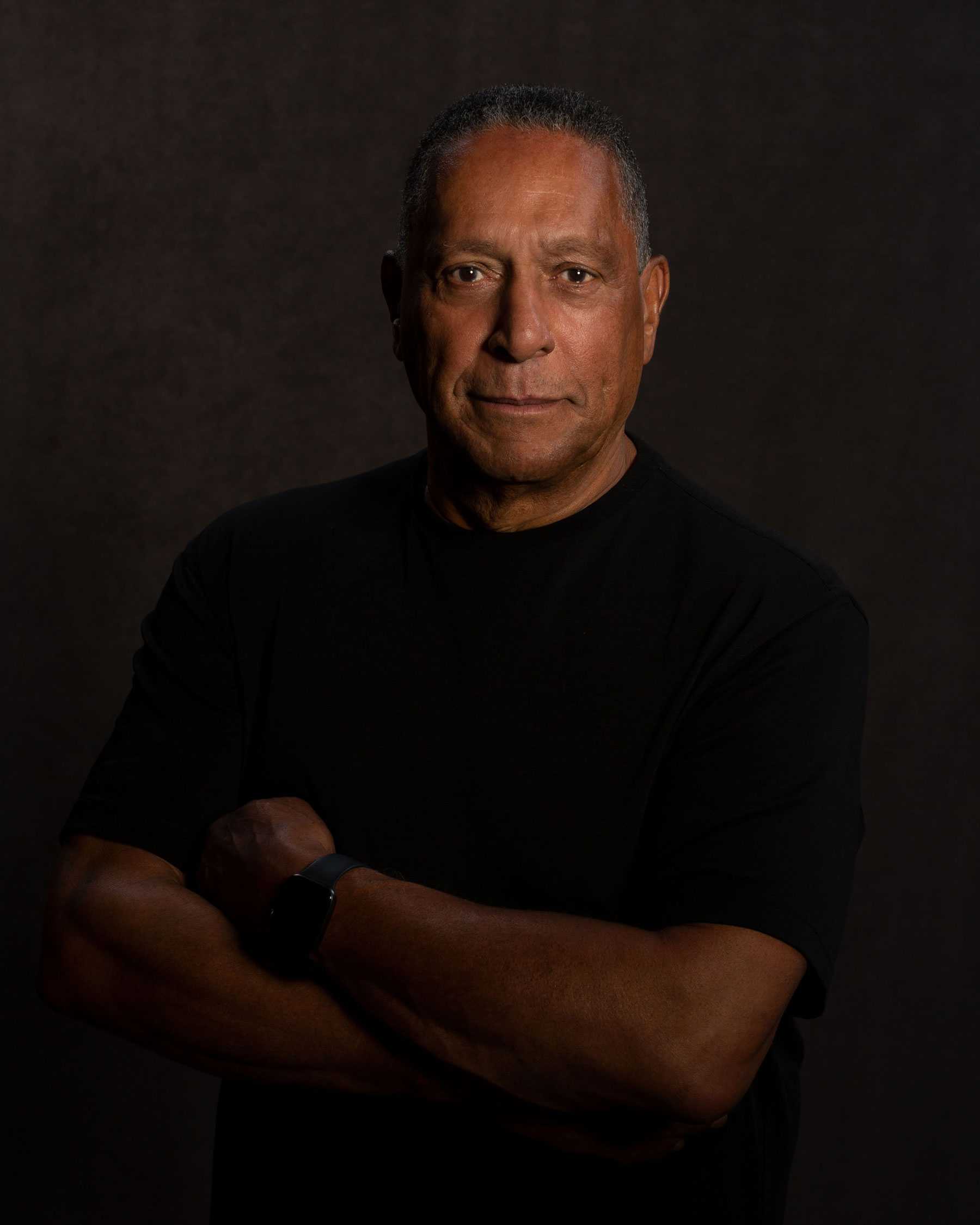 Man in a black shirt poses with arms crossed against a dark background.