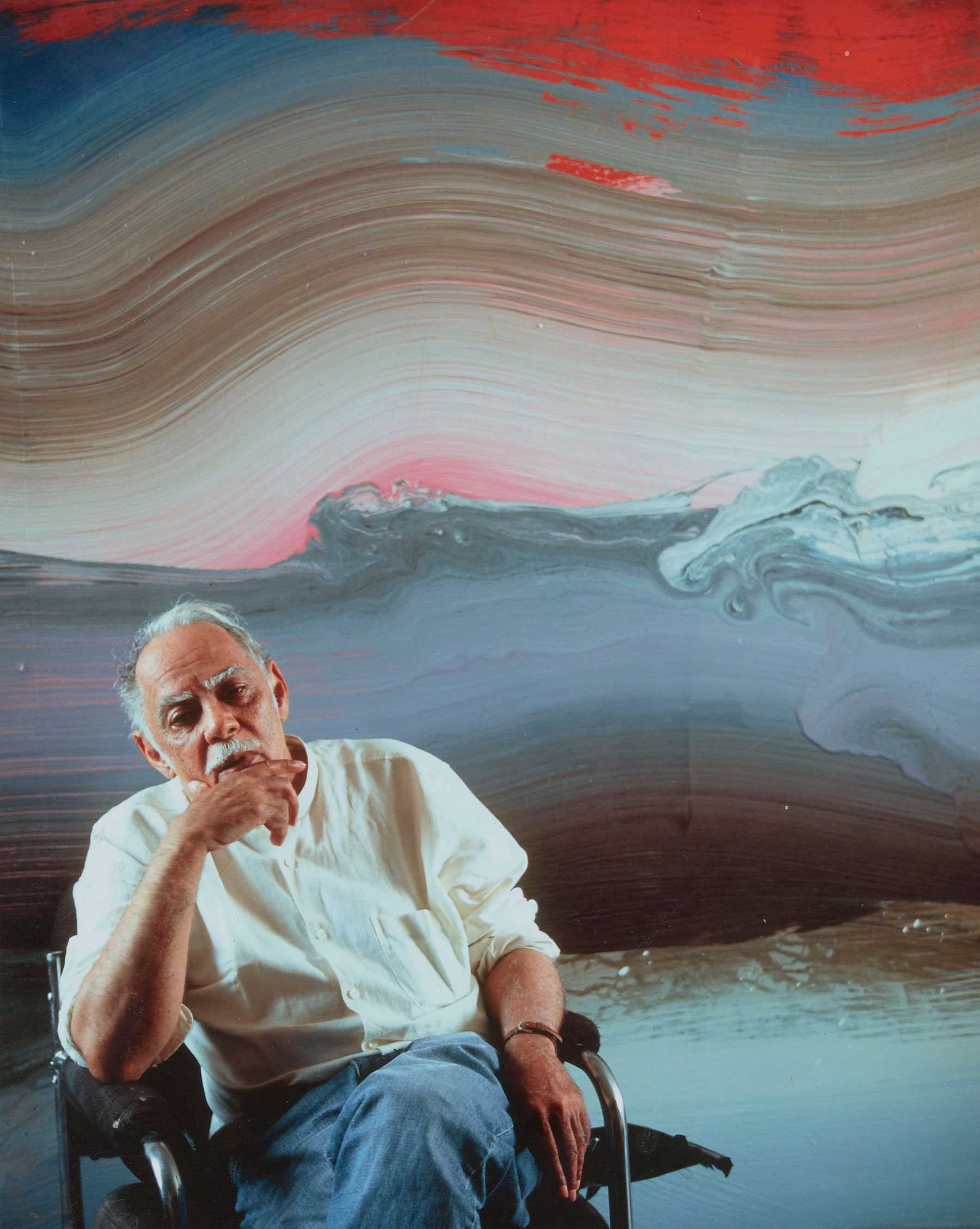 Man seated in front of a colorful abstract painting, wearing a white shirt and blue jeans, hand on chin.