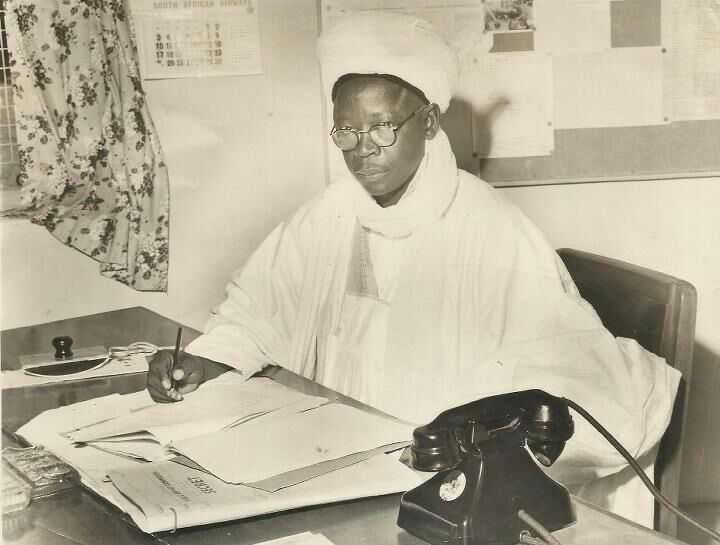 A photo of Abubakar Imam sitting at a desk in an office. He is writing on some papers and a newspaper underneth.