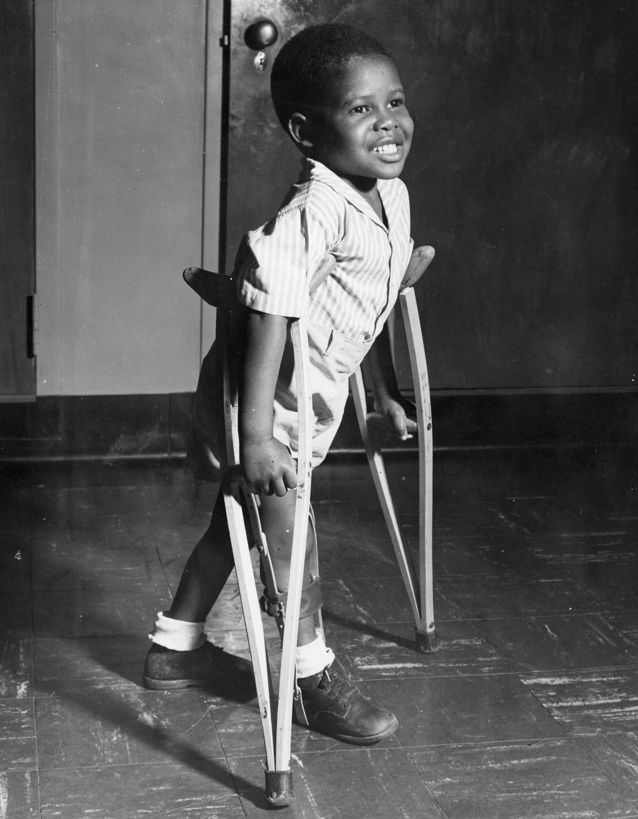 A young child smiling and using crutches, standing on a polished floor, wearing a striped shirt and shorts.