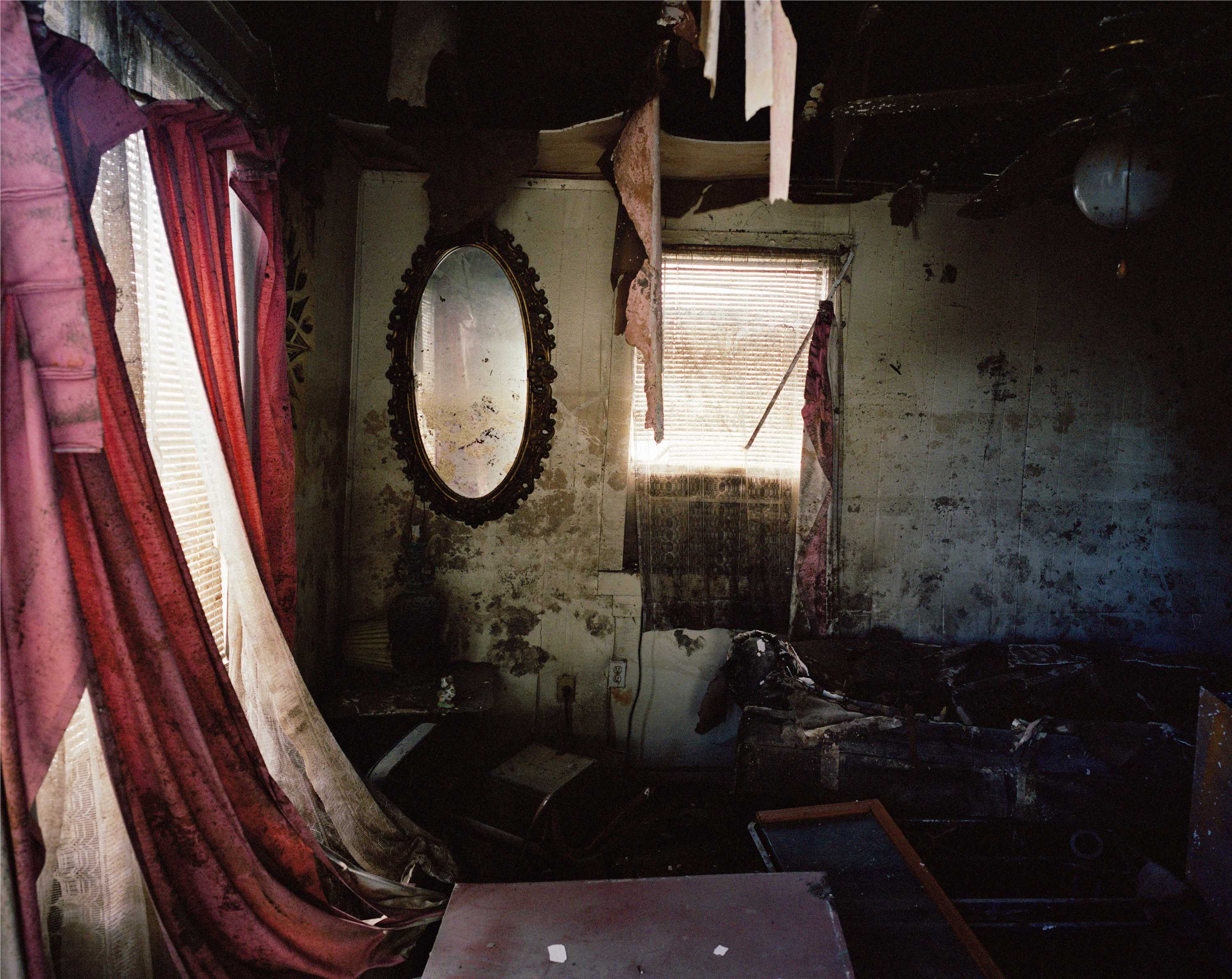 Color photograph of an interior room damaged by Hurricane Katrina. To the left are windows obscured by red drapes and blinds. On the far wall is an oval mirror and another window. The walls are covered in mold. The floor is covered in assorted debris, and parts of the ceiling have fallen or are dangling.