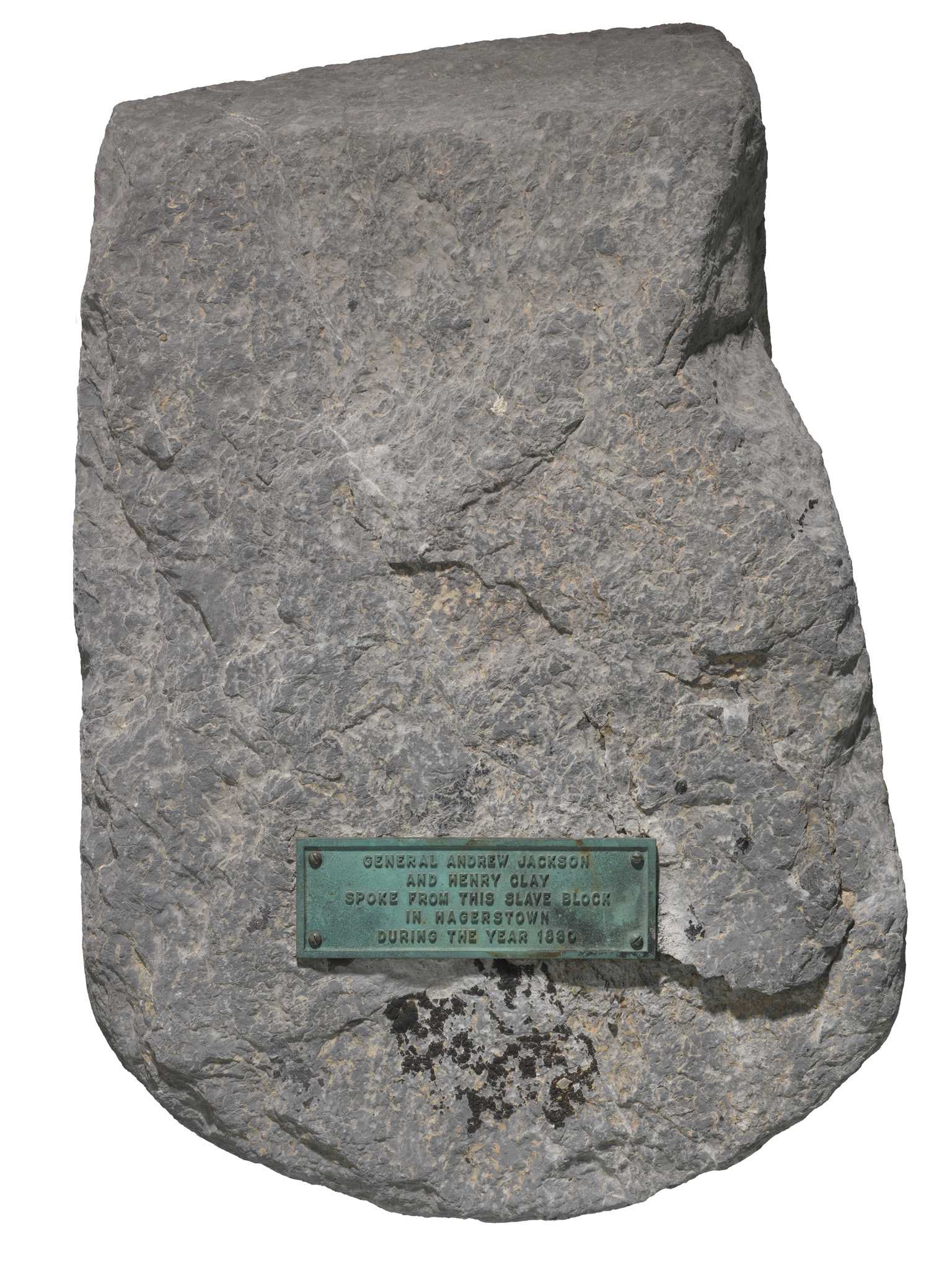 A large gray carved marble Calc-silicate shist stone with a flattened top and bottom, squared back and sides, and a rounded front used as a slave auction block in Hagerstown, Maryland. A rectangular metal plaque is screwed to the top of the stone, with embossed text reading “GENERAL ANDREW JACKSON / AND HENRY CLAY / SPOKE FROM THIS SLAVE BLOCK / IN HAGERSTOWN / DURING THE YEAR 1830.”