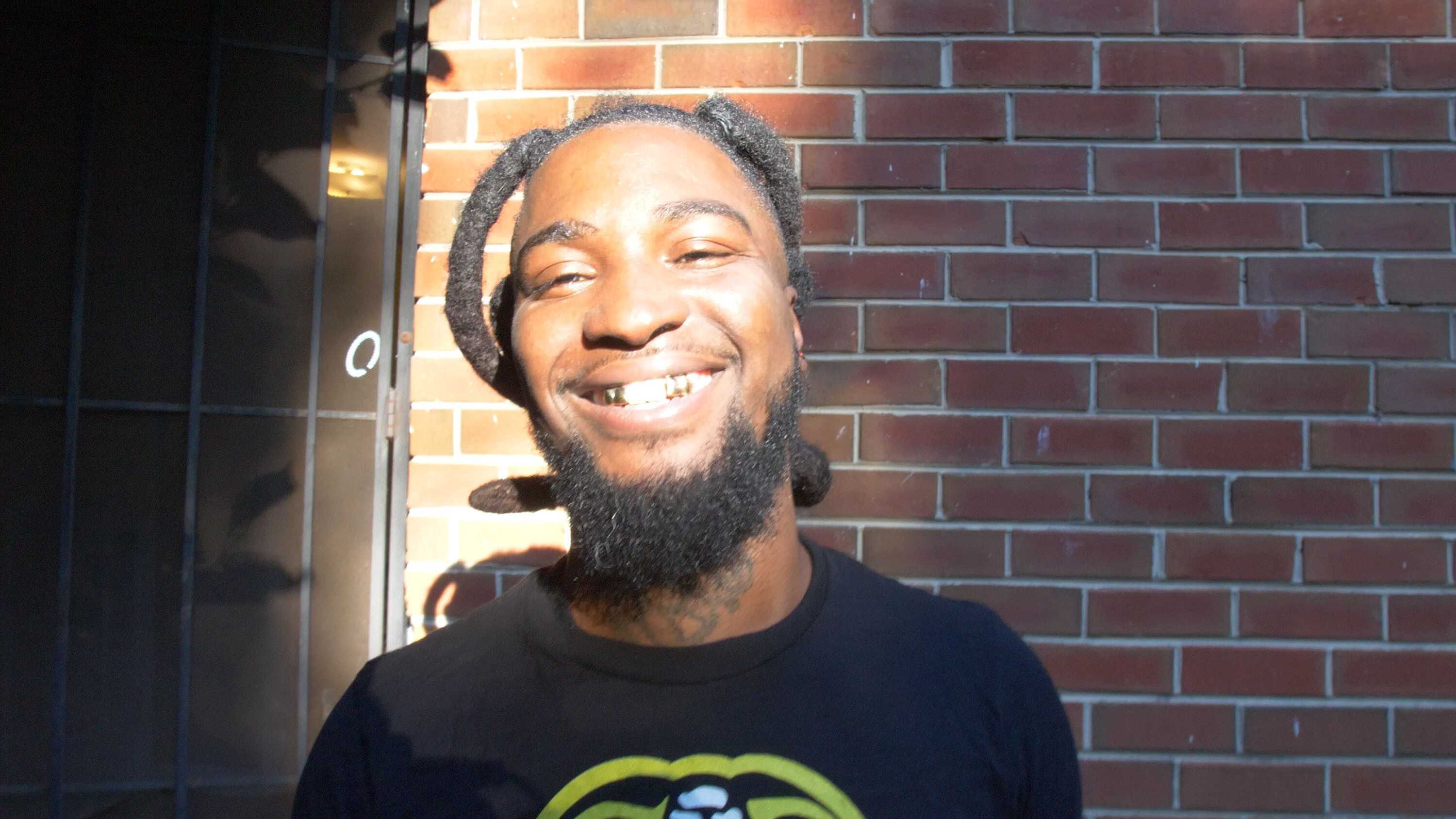 Close-up outdoor portrait of a person with braided hair and a beard, standing in bright sunlight against a red brick wall, wearing a dark T-shirt.