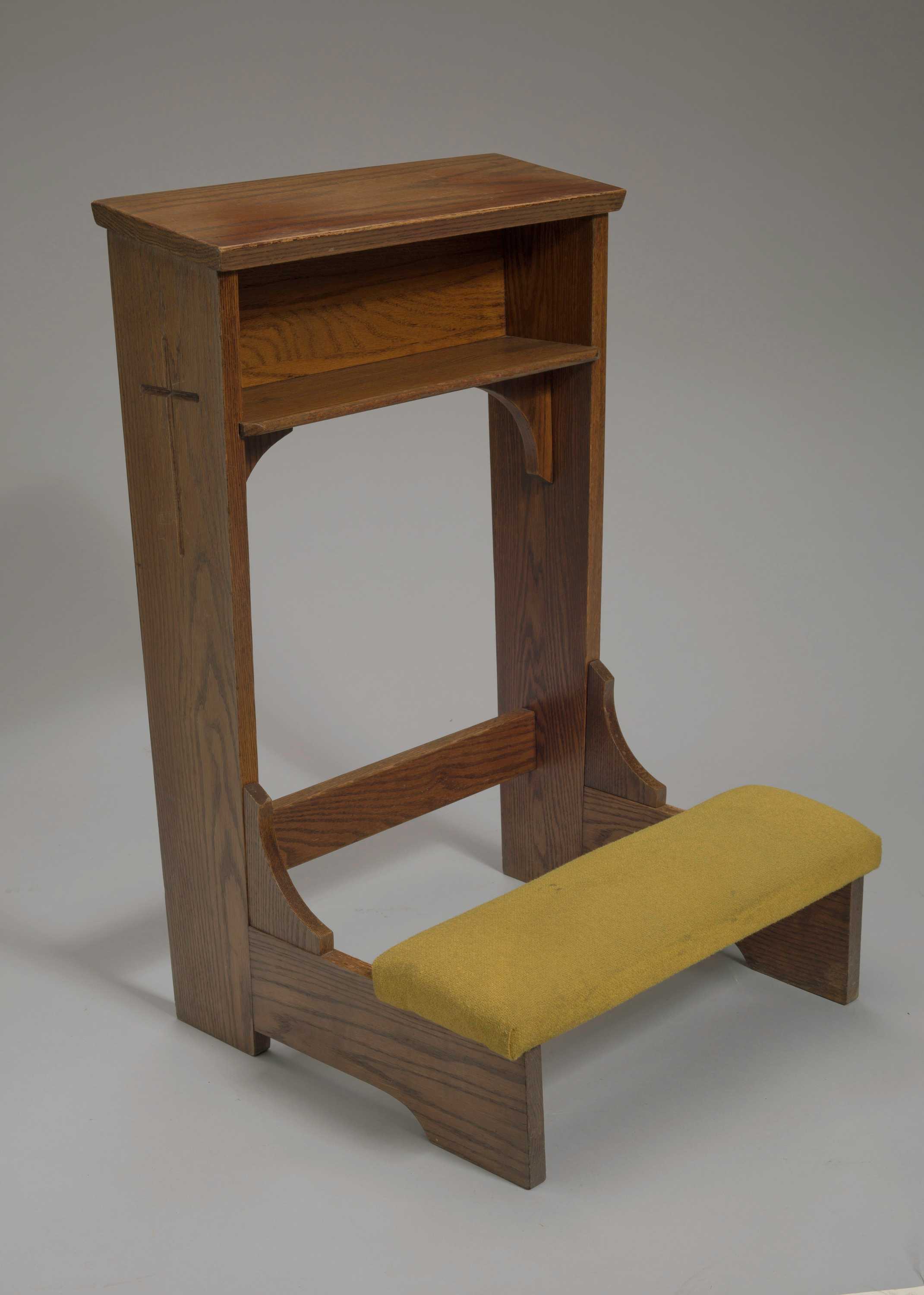 A wood kneeling altar with a yellow-green padded knee rest. The altar has small cubbyhole under the top shelf. The shelf is slightly tilted down toward the knee pads. The bottom of the shelf and cubbyhole are supported by two curved arches on either side, attached to the side planks. On either side of the altar at the top of the side planks is a carved cross. A small rectangular support board is near the base of the altar and connects the two side planks. Kneepads are attached to the fixed base with two solid horizontal planks on either side. A small, wood, inverted arch support connects the knee rest to the vertical side planks.