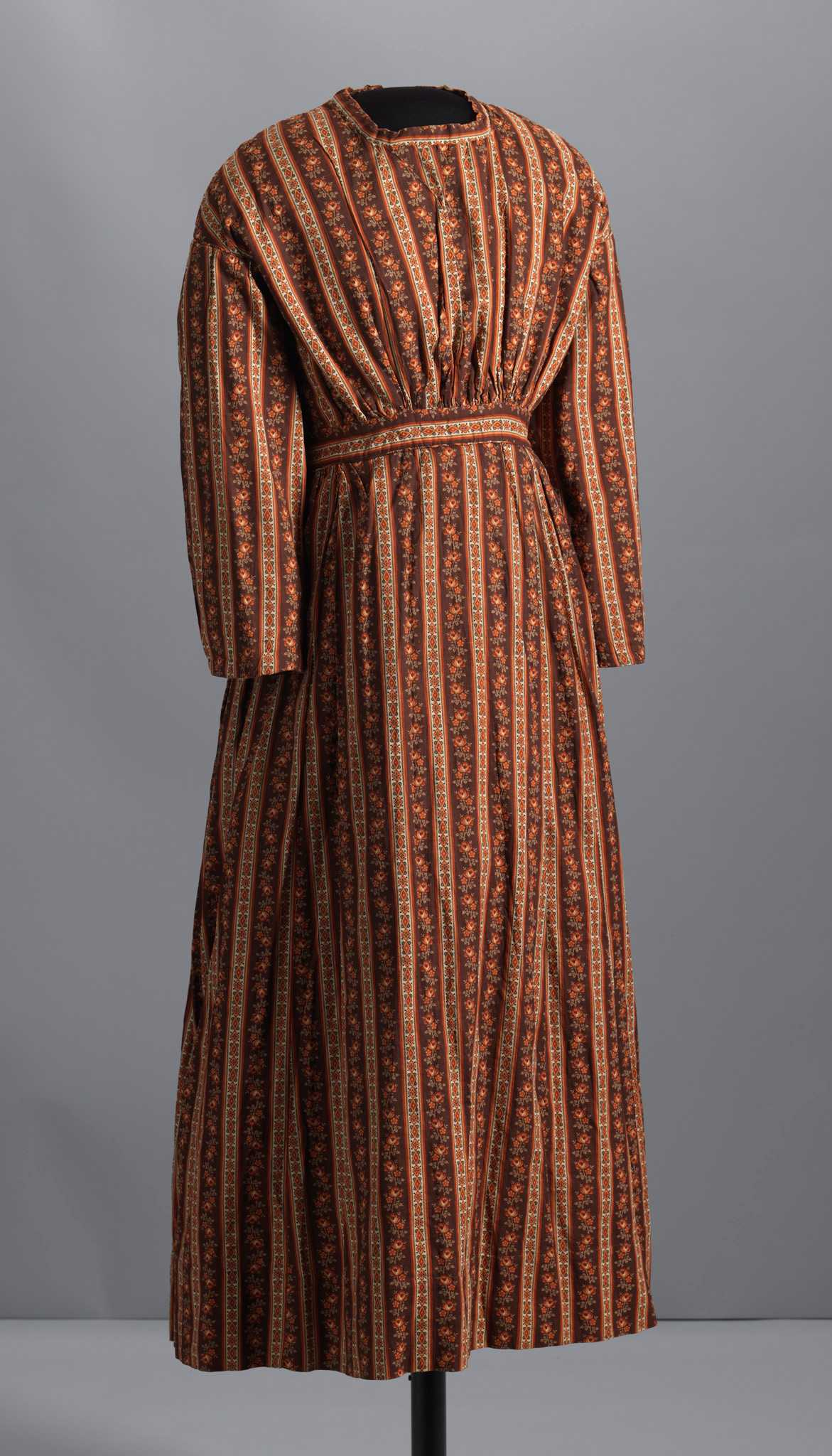 This is a handstitched long-sleeved dress with a full skirt made from a balanced plain-weave printed cotton. The sleeves are gathered at the back shoulder seam and the shoulder seam is dropped in the back. The bodice is gathered at the front waist and has a neck band with no collar. Both the neck band and the waist band are fussy cut to display the printed design. Piping is attached at the armholes and along the bottom of the waistband. The skirt is gathered at the front hips and the back with a flat panel along the center front waist. On the proper back bodice, three (3) glass buttons are sewn on the left side of the back opening (placket), with four (4) button holes on the right side. At the waist, two (2) decorative buttons are sewn on the right side of the opening, without corresponding button holes on the left side. The hem is turned up 3 1/2". The bodice and sleeves are lined with muslin, while the skirt is unlined. The printed pattern on the fabric is four (4) colors and black, with a base color of reddish brown, on top of which is a repeating pattern of columns of red roses alternating with columns of diamonds and clubs on a cream background inside a black, orange, and red border.