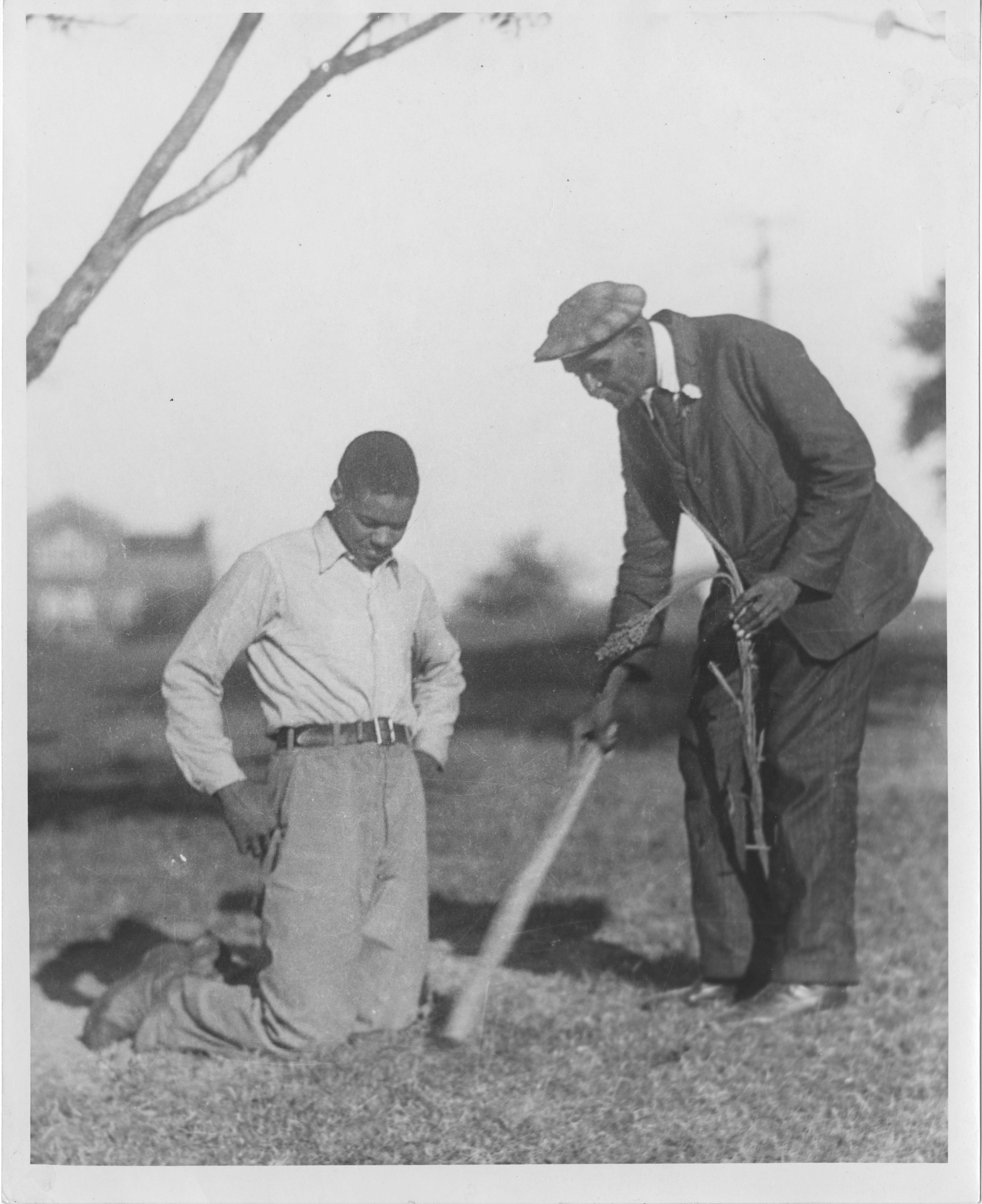 Black and white photograph of George Washington Carver holding a plant while showing a much younger man something in the ground.
