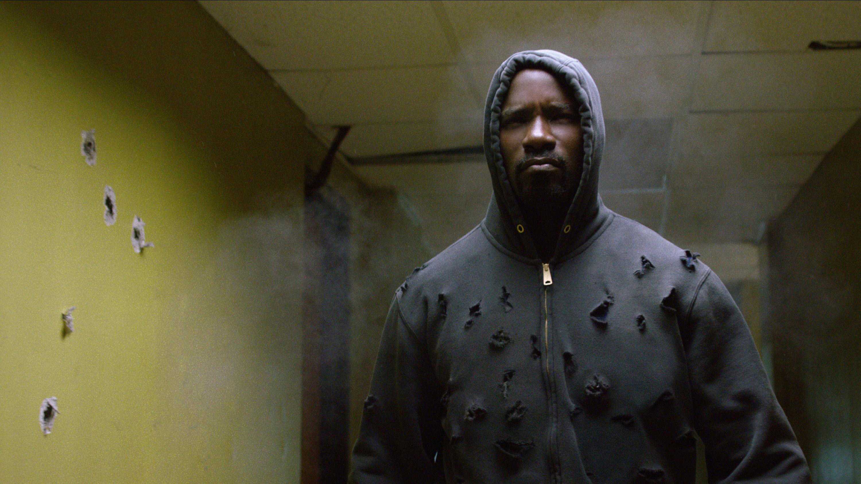 Mike Colter, as Luke Cage, walking through a hallway, with many gunshot holes in his black sweatshirt and along the walls.