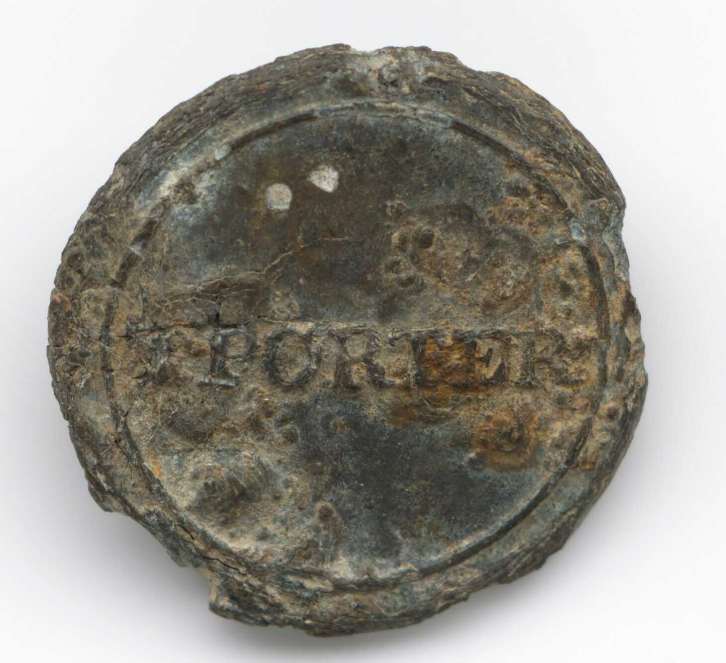 A round pewter button with "TPORTER" stamped across the middle. This button would have been sewn onto an enslaved person's shirt to identify him or her as belonging to Thomas Porter II. On the reverse side of the button there are fine concentric circle impressions within a pronounced rim as well as a stamp which is now illegible. There is also evidence of a parting line for a two-part mold on the reverse of the button. The button has considerable wear with pitting on both sides.