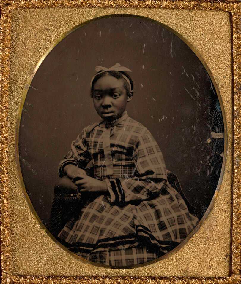 Tintype of young girl sitting in fancy plaid dress with a white bow in her hair. She is leaning to the proper right and resting on the arm of a chair. The tintype is framed in an oval mat with a gold frame. The tintype is in a case with a floral  design on the front and back. The inside of the case, opposite the tintype is velvet with an embossed floral design.