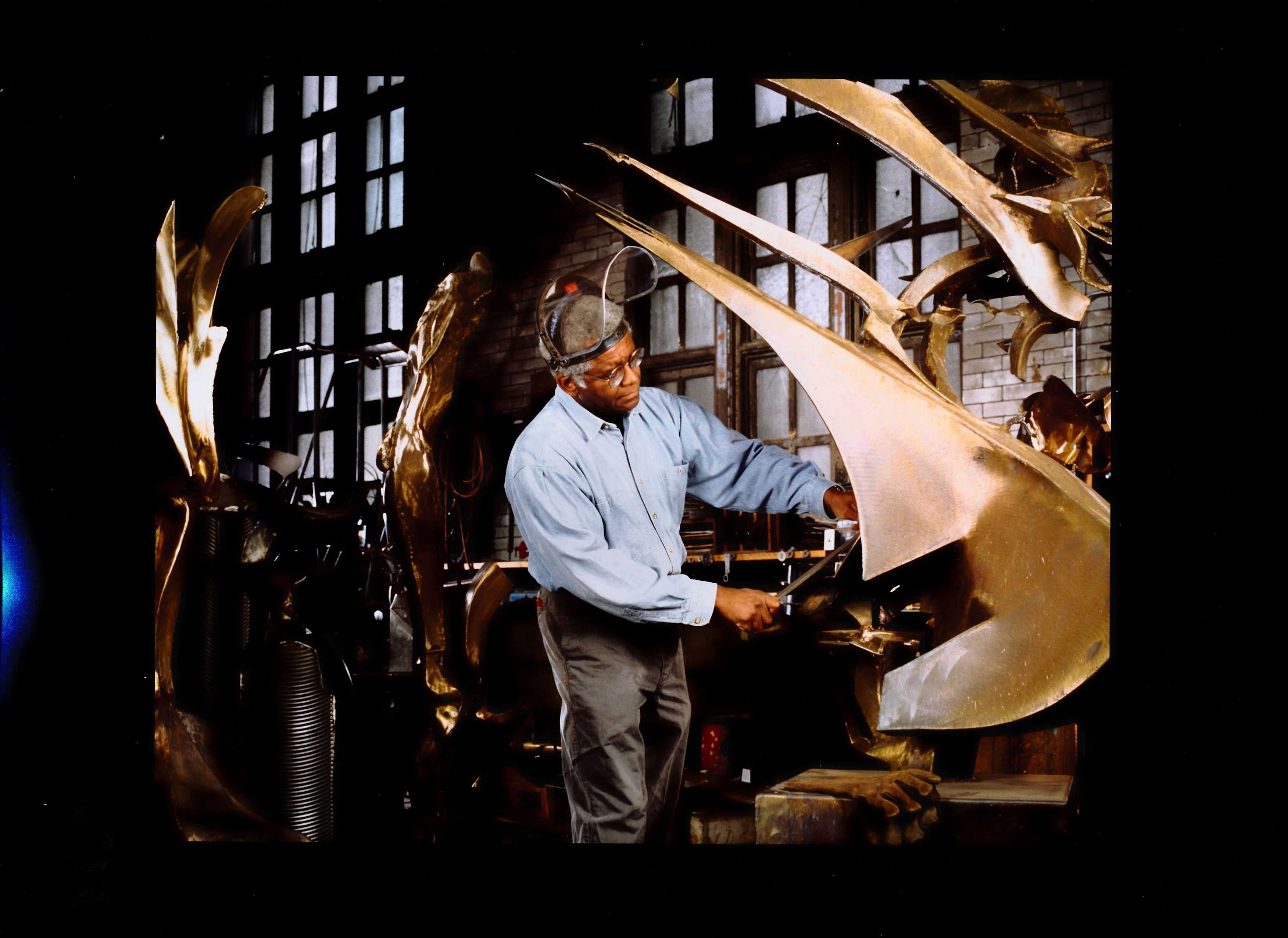 Person wearing a face shield and glasses works on a large abstract metal sculpture in a workshop filled with tools and bronze pieces.