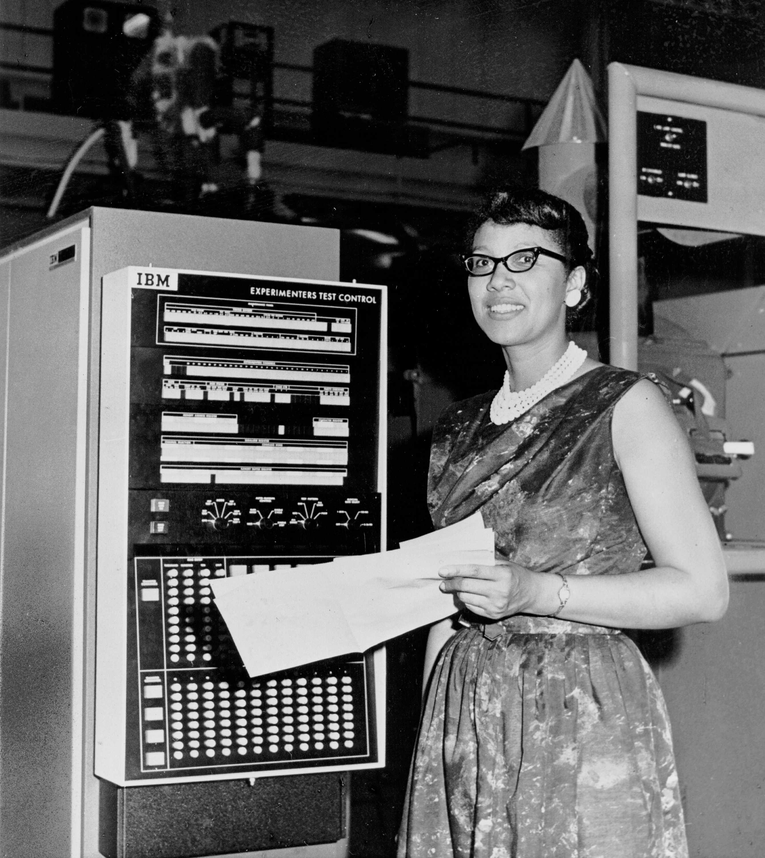 Melba Roy Mouten is holding paper in front of some machines as she smiling for a black and white photograph