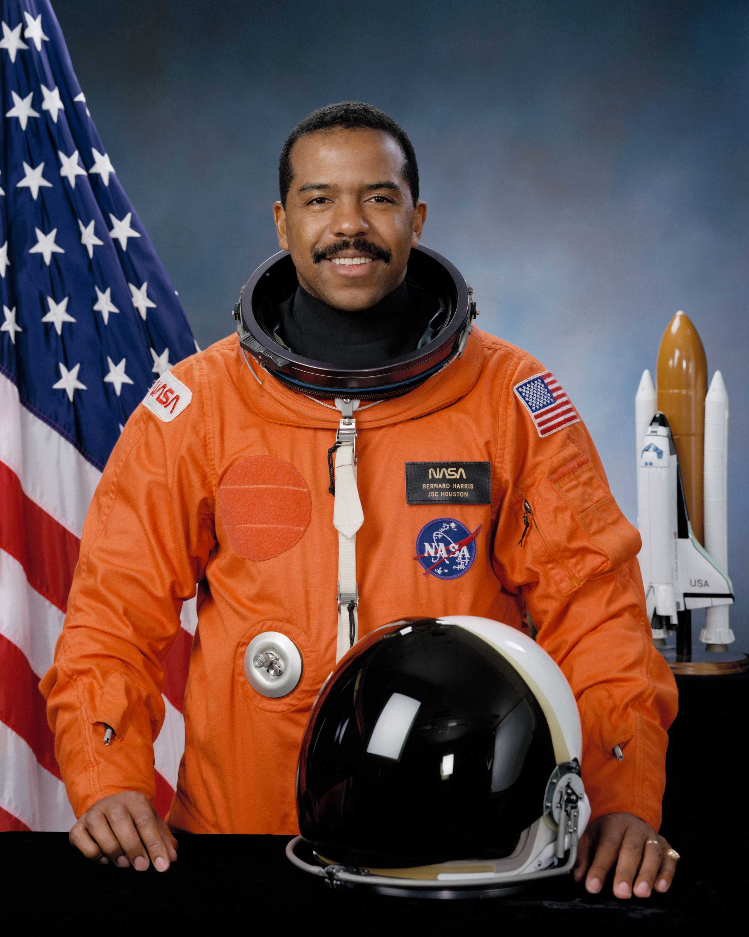 Harris is wearing an orange space suit with a helmet resting on the table in front of him while he smiles for his portrait.