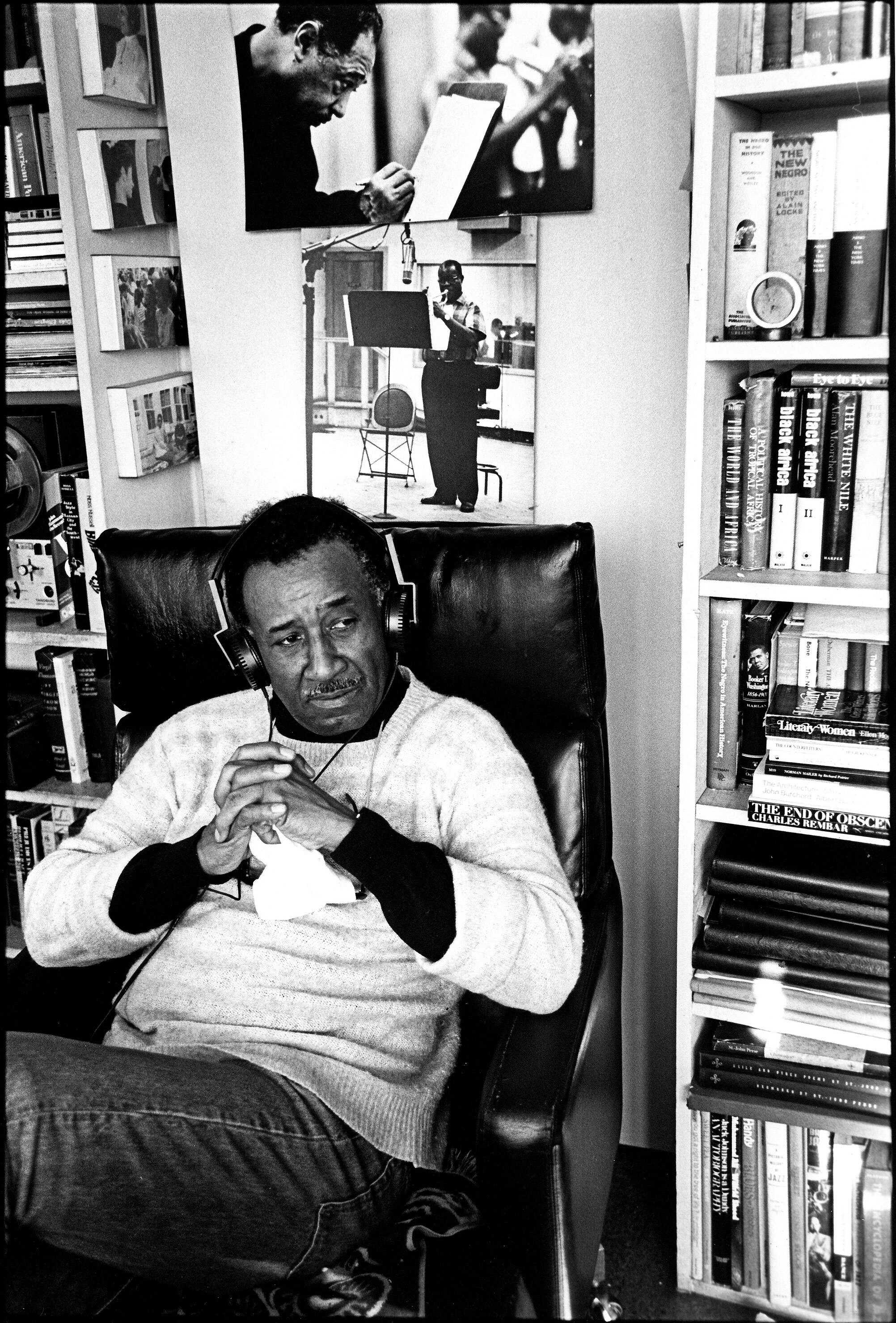 Person seated in a chair in a room lined with bookshelves, with two black-and-white photographs displayed on the wall behind.