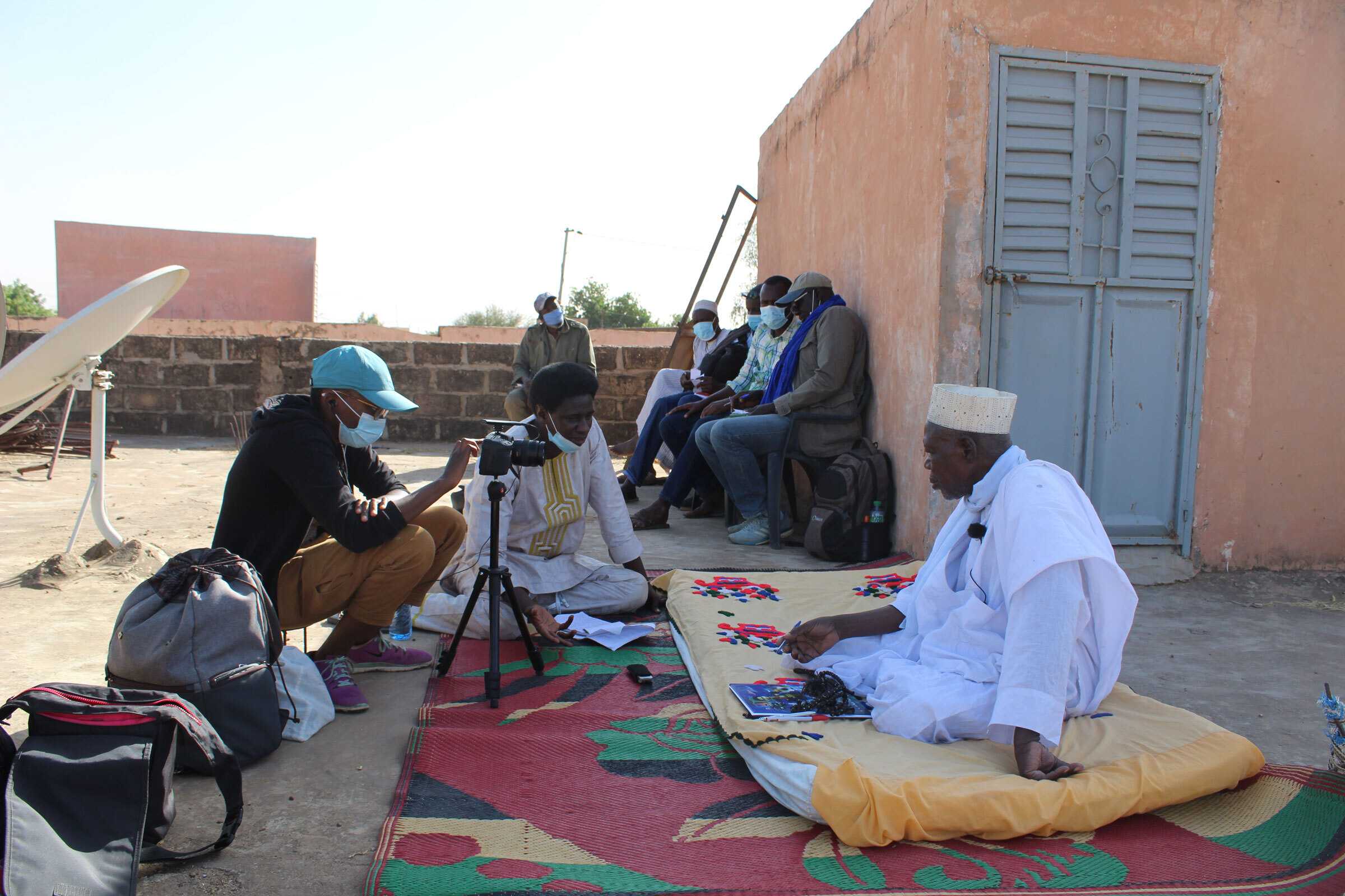Outdoor interview or documentation session beside a building, with one person seated on a mat demonstrating colorful pieces on a cloth while another records using a camera on a tripod; several people sit and observe in the background.