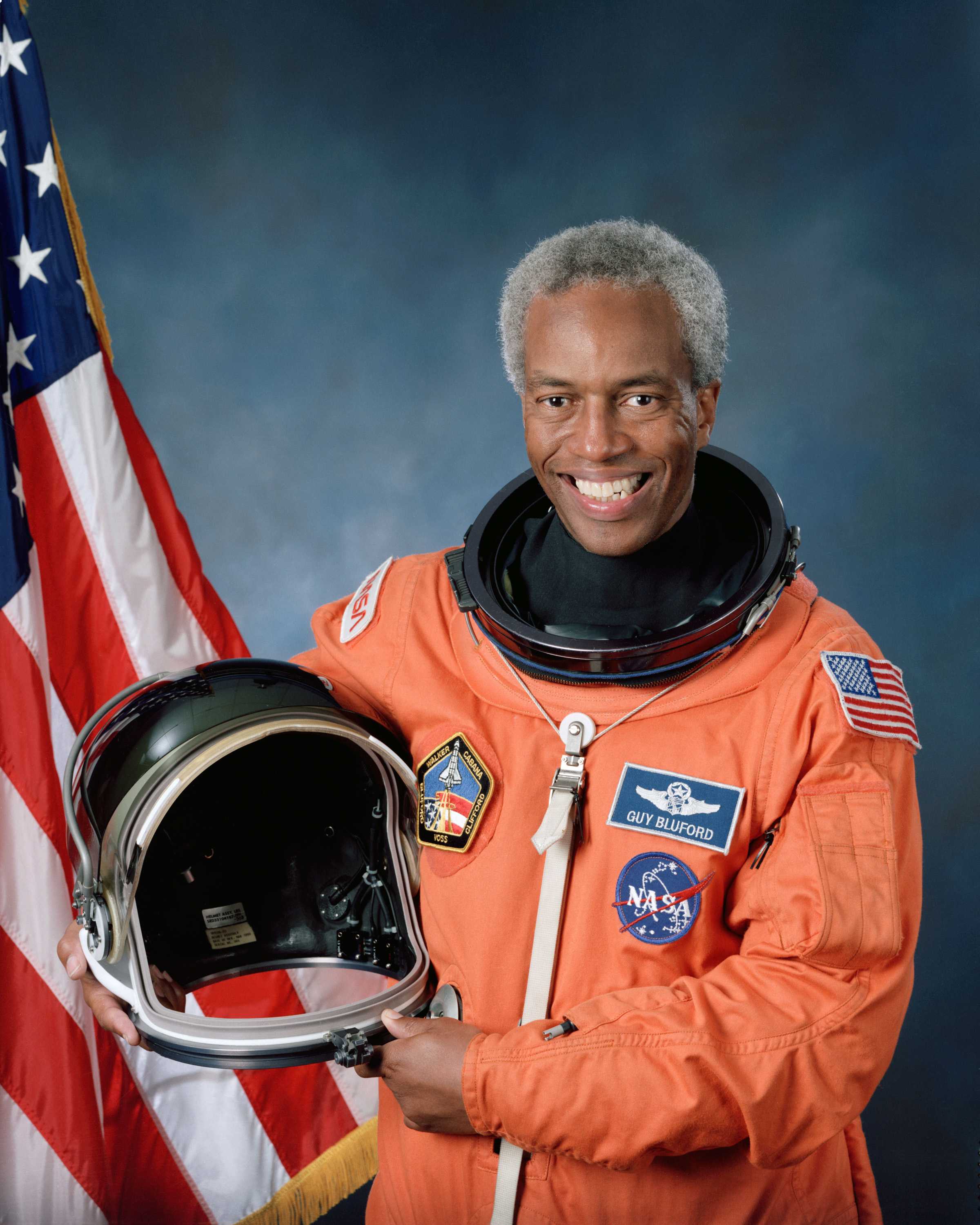 Guion Bluford poses for a portrait in an orange NASA space suit, holding a helmet, in front of a blue background.
