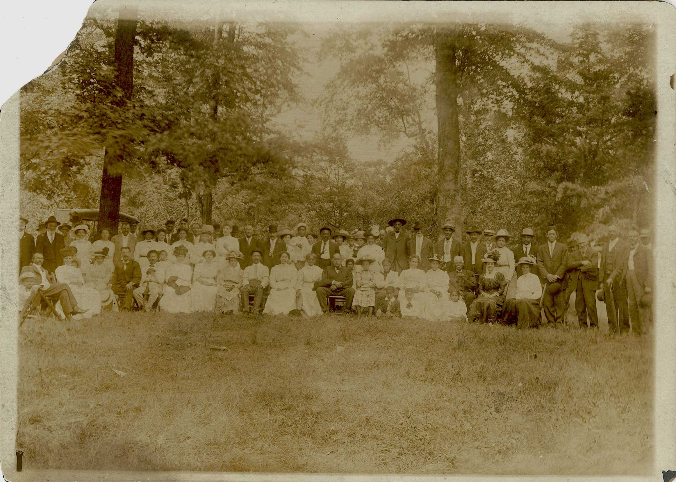 A sepia tone photograph of a family reunion. About 30 people are posed for the portrait in a wooden area.