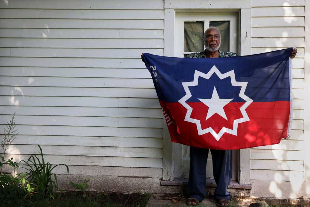 Person standing in a doorway of a light-colored house holds a red, white, and blue flag with a large starburst emblem.
