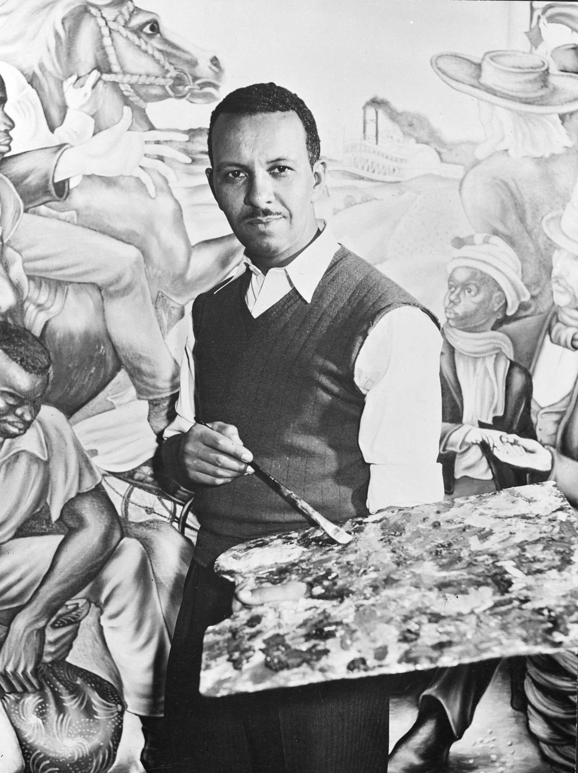 Black-and-white photo of an artist holding a palette in front of a detailed mural depicting various figures and scenes.