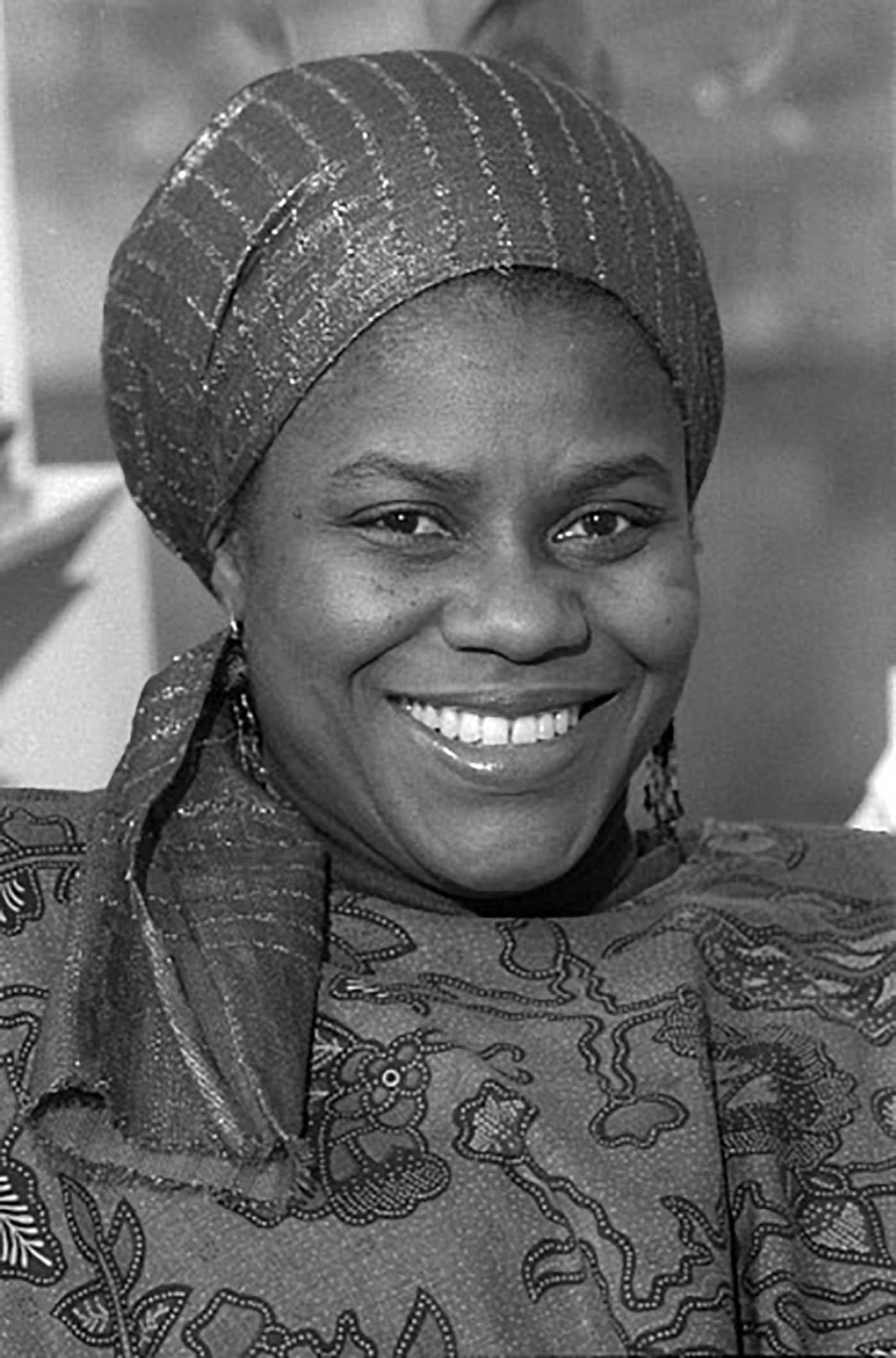 Black-and-white photo of a smiling person wearing a patterned headscarf and garment.