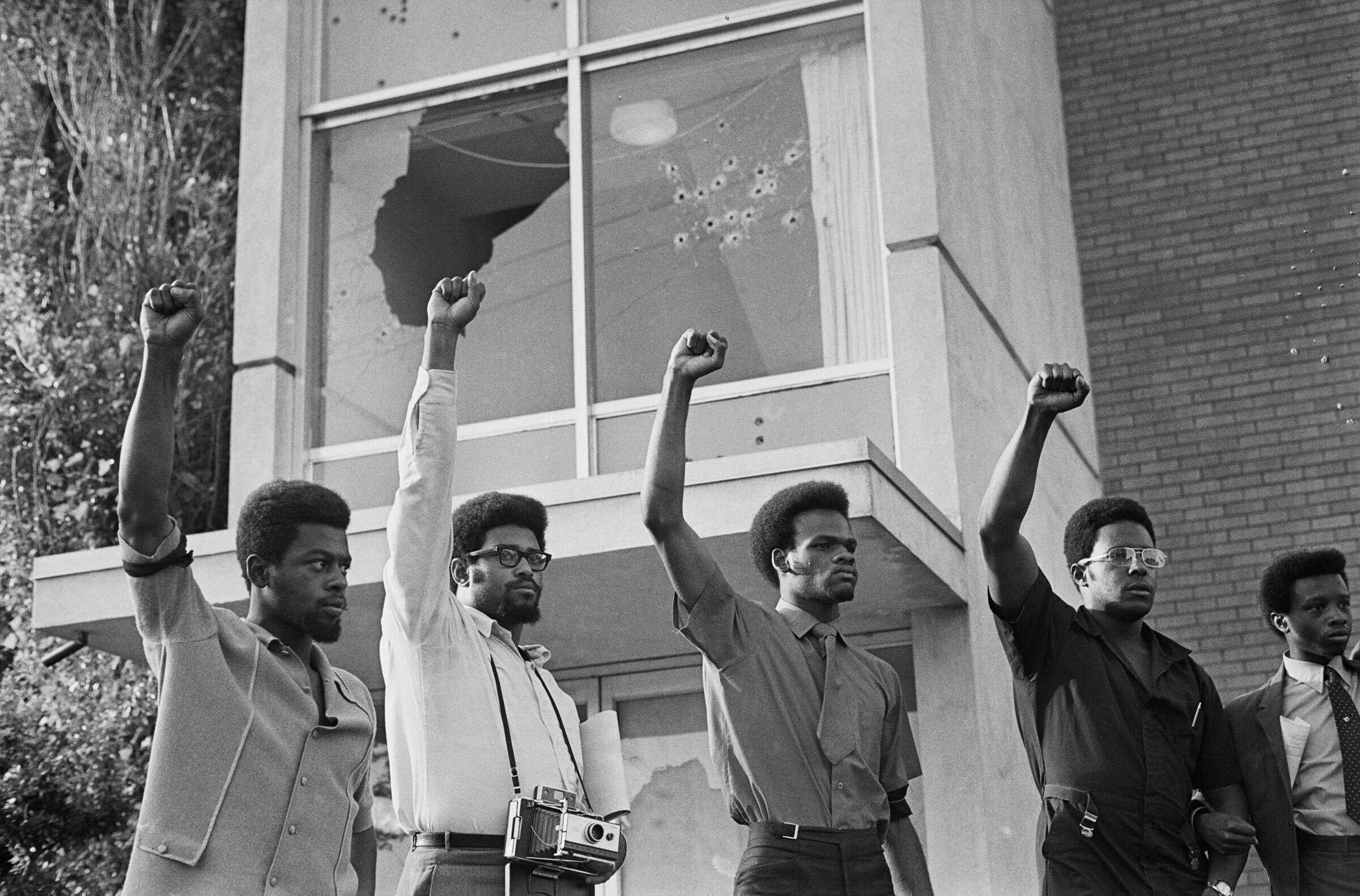 People standing in front a building with a broken and damaged window.