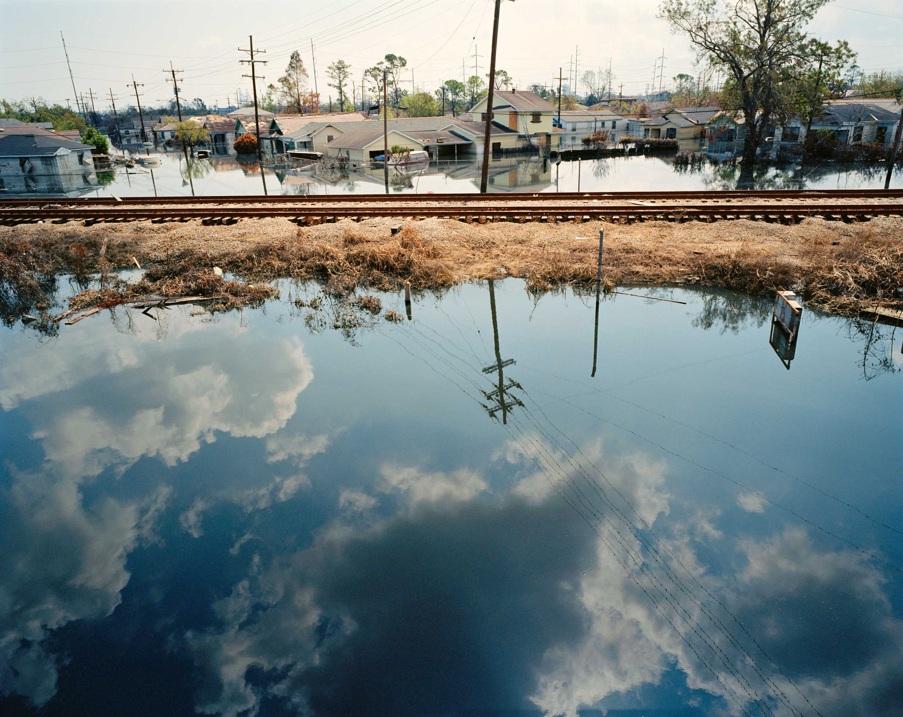 A color photograph of flooding in New Orleans in the aftermath of Hurricane Katrina. In the foreground is calm floodwater reflecting a partly cloudy sky. Behind the reflection is a railroad.