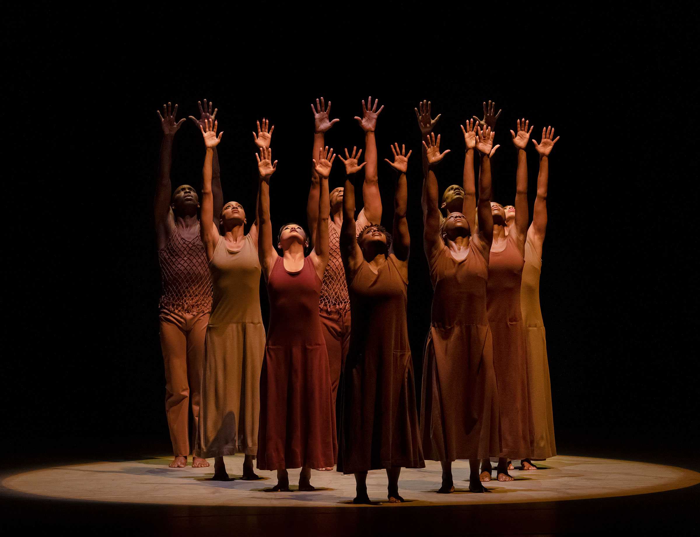 A group of dancers on stage with arms raised, wearing earth-toned costumes, performing under spotlight in a dark setting.