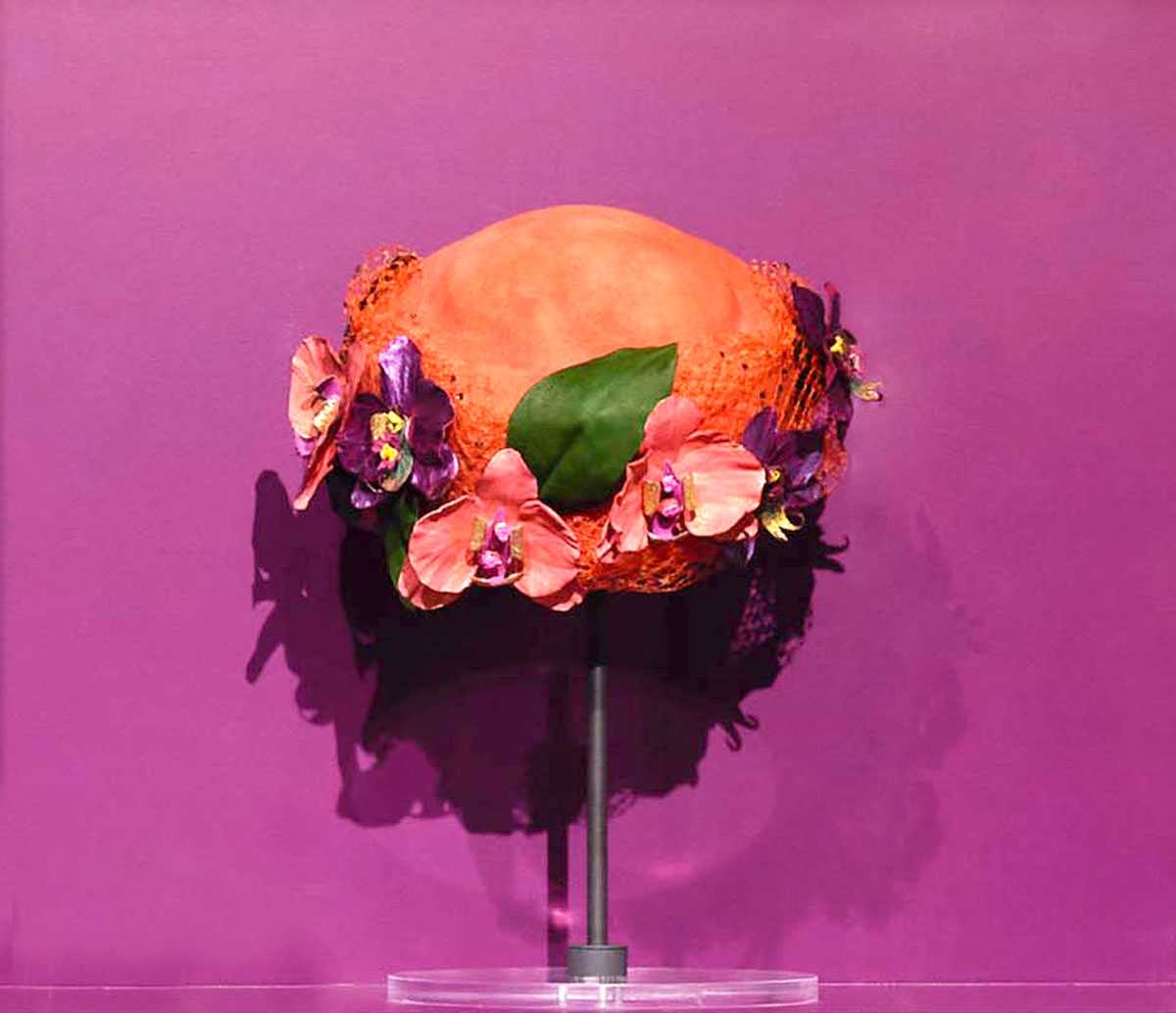 Ornate hat adorned with pink and purple flowers, displayed on a stand against a purple background.