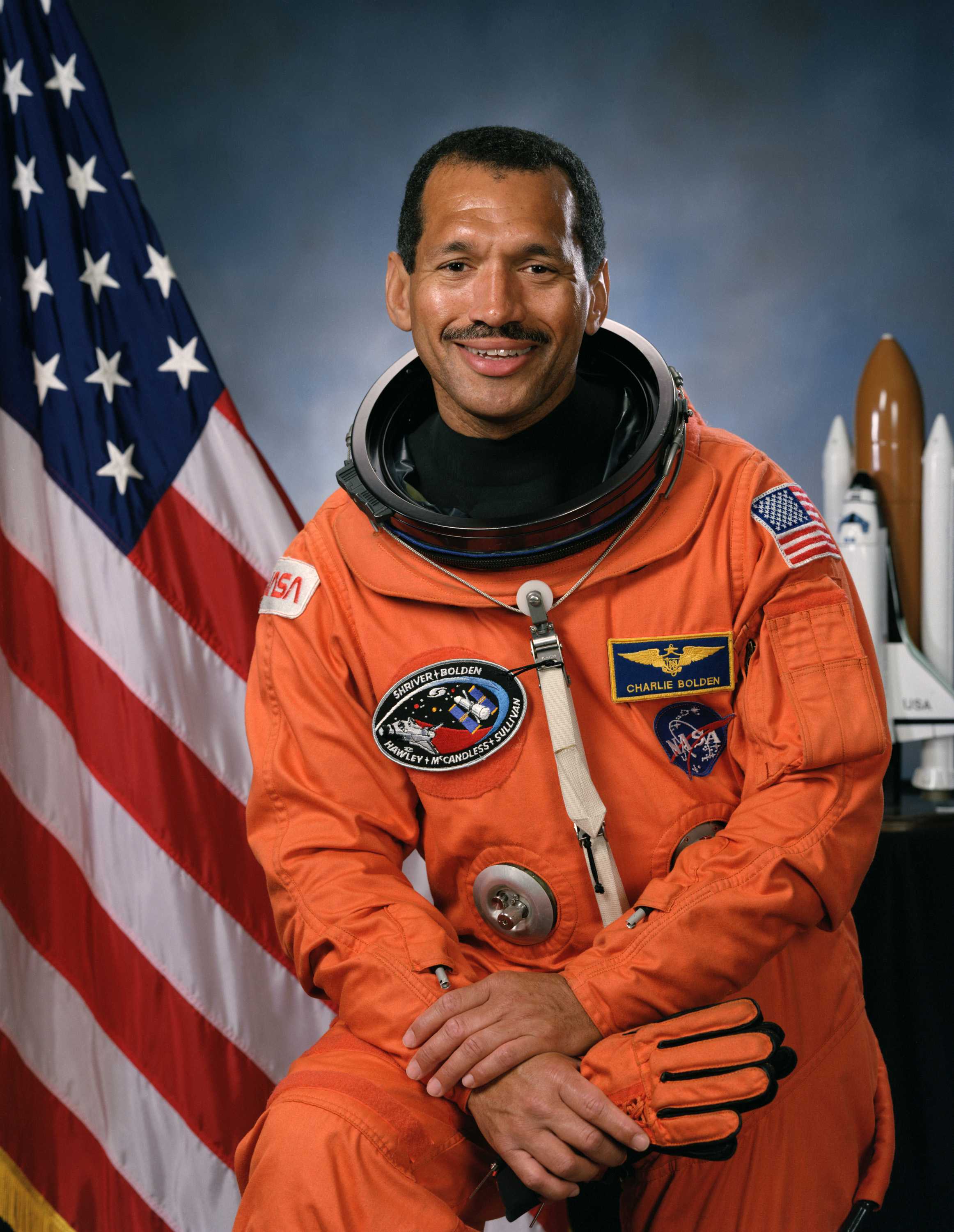 Bolden is wearing an orange space suit while sitting and smiling for his portrait.