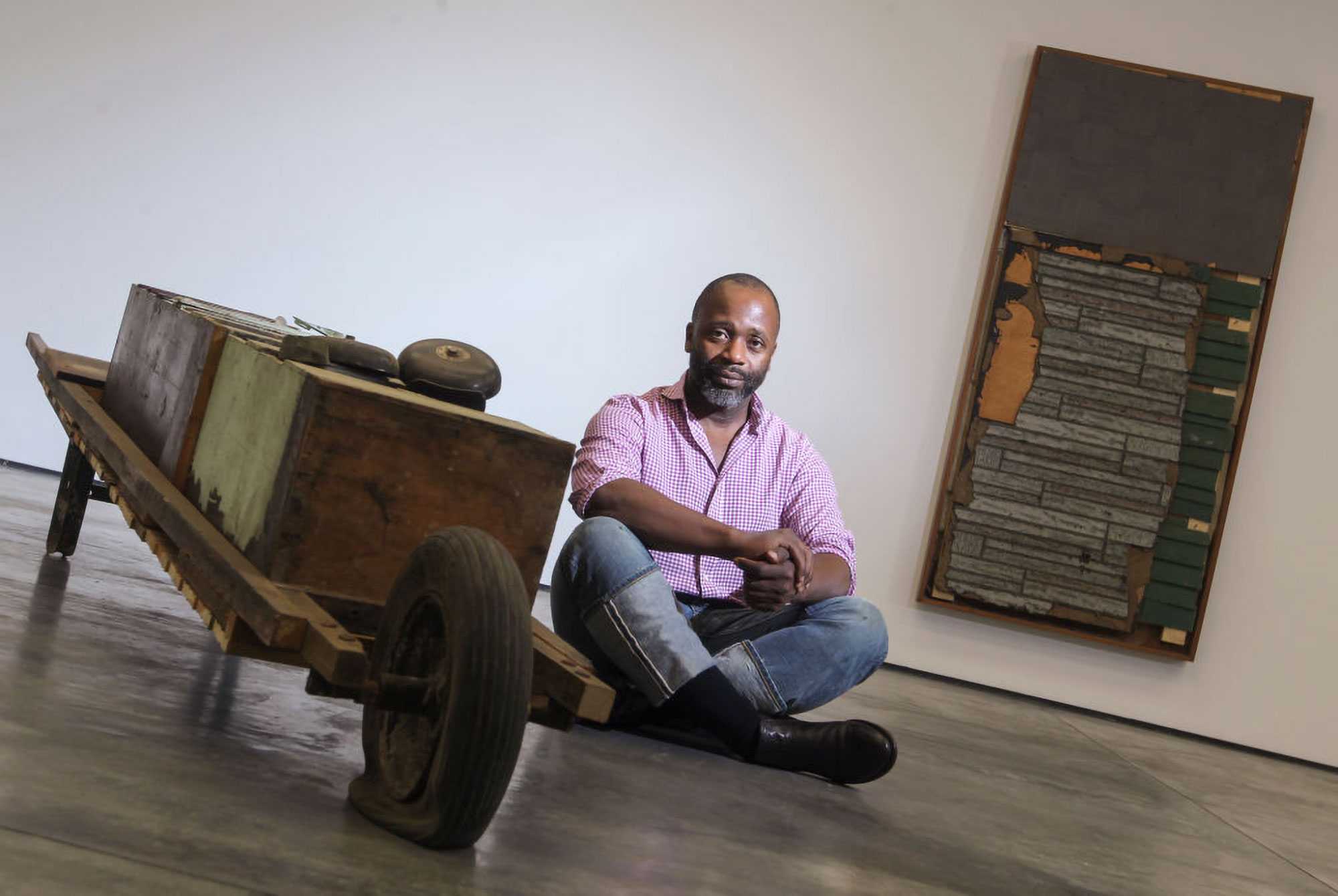 Man sitting on the floor near wooden cart and artwork leaning against the wall in an art gallery.