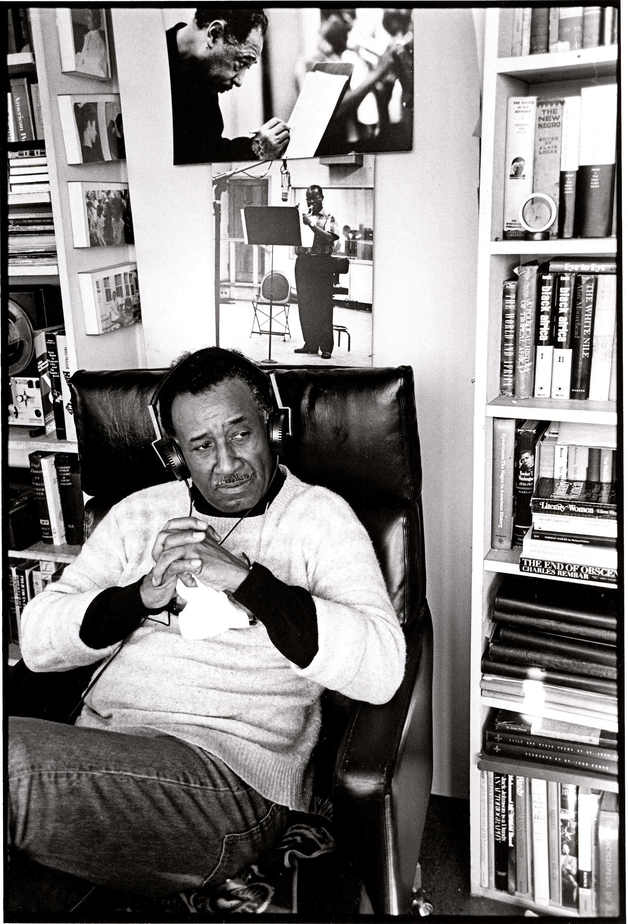 Person seated in a chair in a room lined with bookshelves, with two black-and-white photographs displayed on the wall behind.
