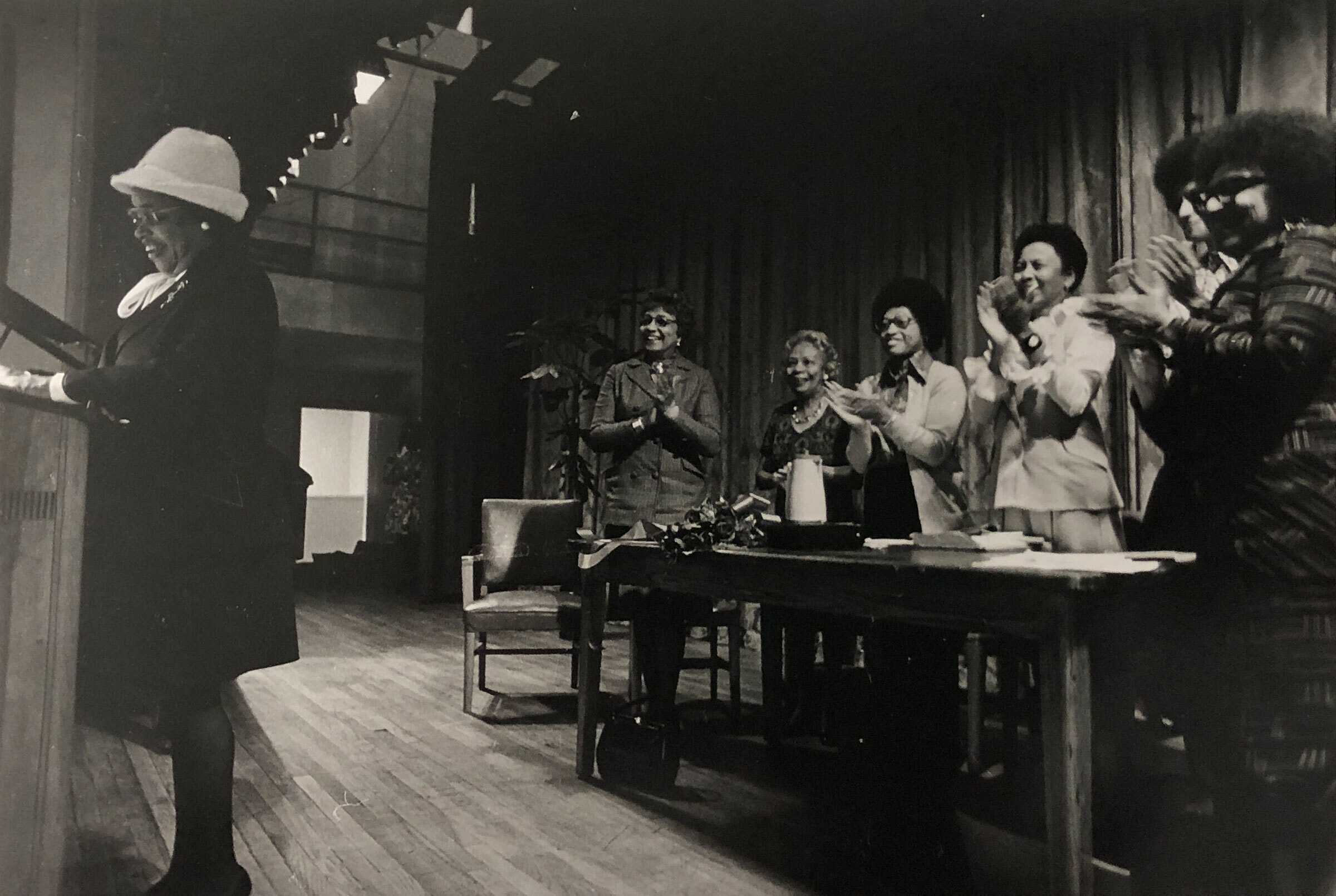 Black-and-white photo of a person standing at a podium on a stage while several people at a long table applaud. The setting includes wooden flooring, a curtain backdrop, and chairs arranged behind the table.