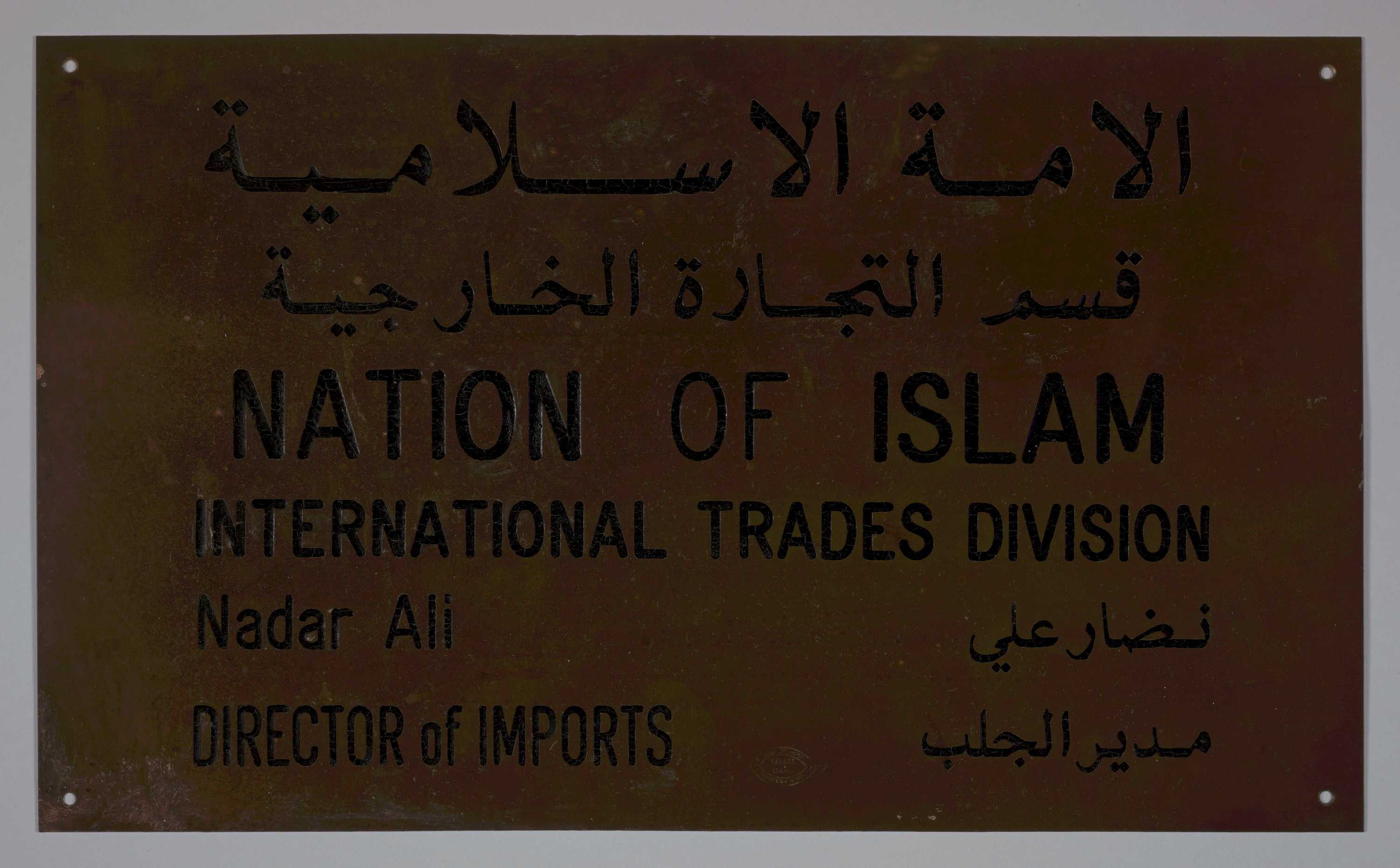 Rectangular, brass sign with four (4) holes, one (1) in each corner. Along the top are two (2) lines of Arabic.