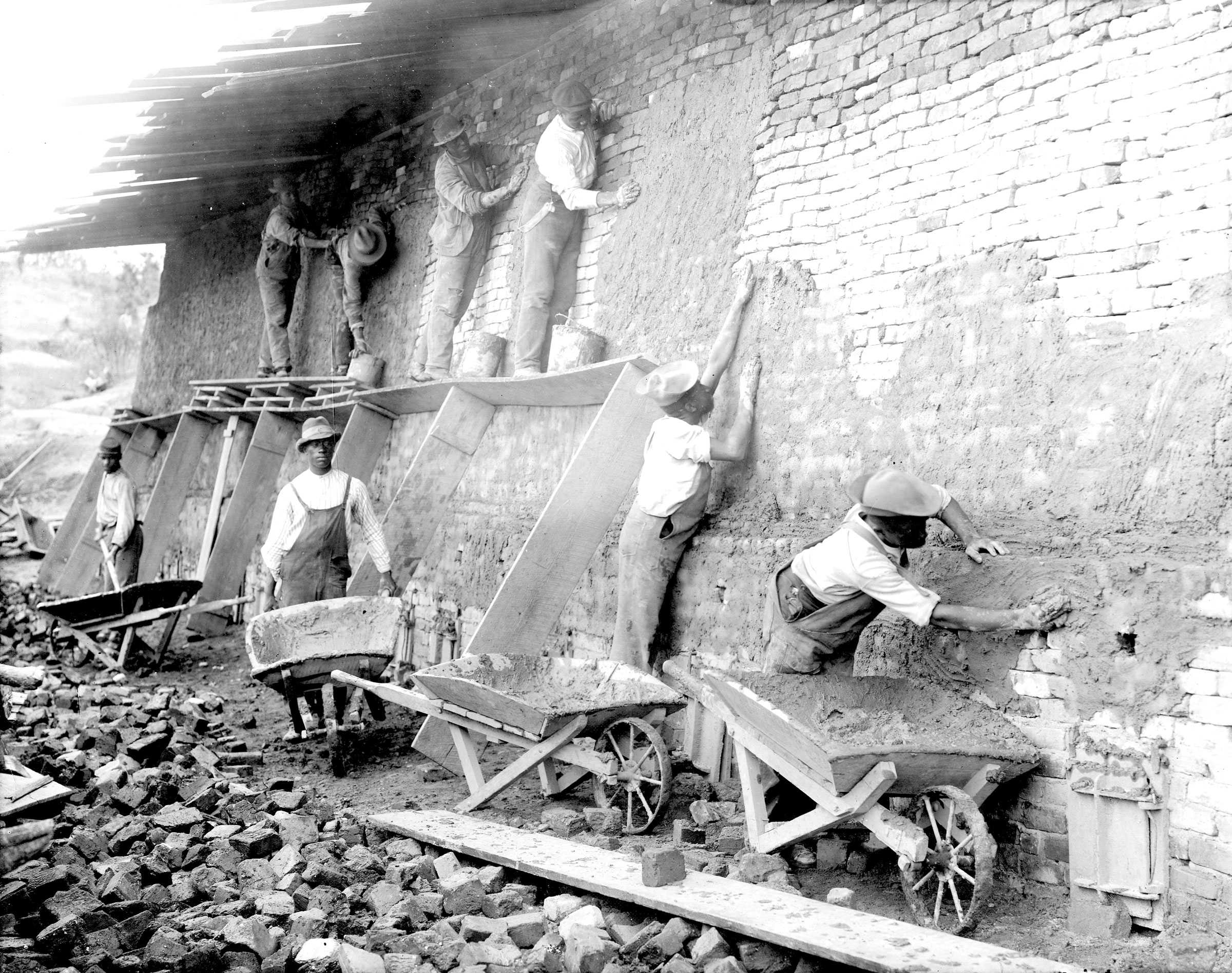 Photo of workers applying stucco to a brick wall, using wheelbarrows and wooden boards for support.