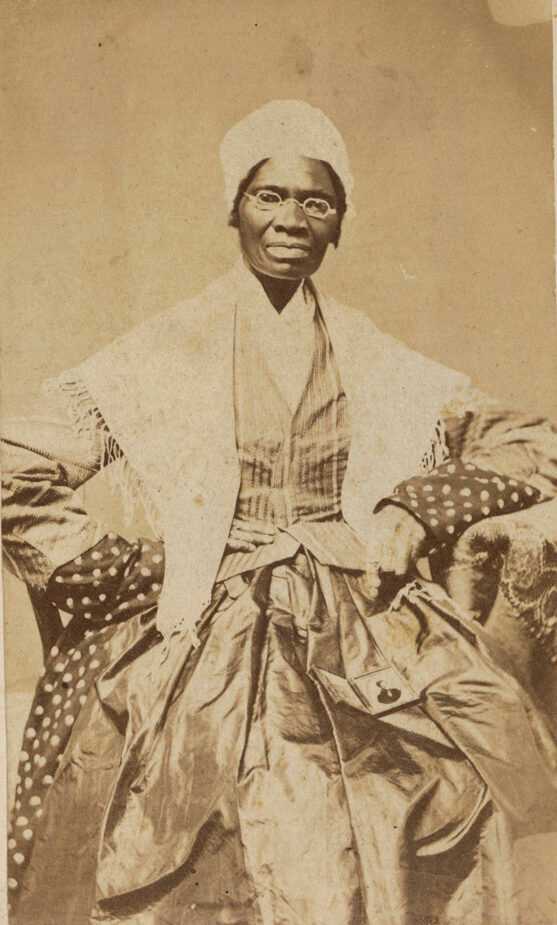 A carte-de-visite portrait of Sojourner Truth, showing the subject seated with a daguerreotype of her grandson, James Caldwell of Co. H, 54th Massachusetts Infantry Regiment, on her lap. Truth wears a dot print dress, striped jacket, dark-colored apron, a light-colored shawl over her shoulders, and a light-colored cap. She looks straight at the camera. Her proper right hand sits at her waist and her proper left hand appears to point at the daguerrotype lying open on her lap. There are no inscriptions on the front. On the back is the inscription [Sojourner Truth] and an identification number.