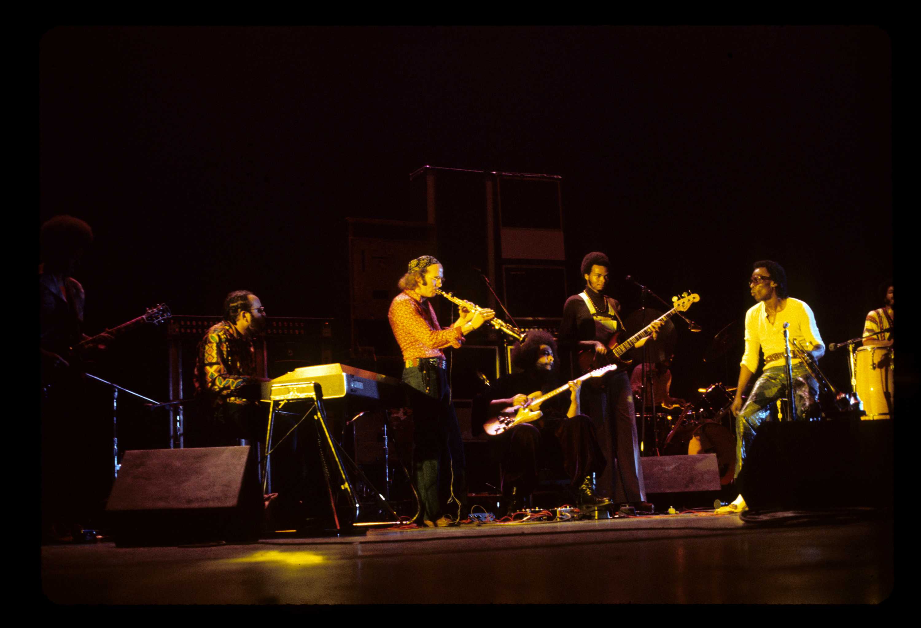 A concert view of Mile Davis on stage with a full band, lite by a warm yellow light.