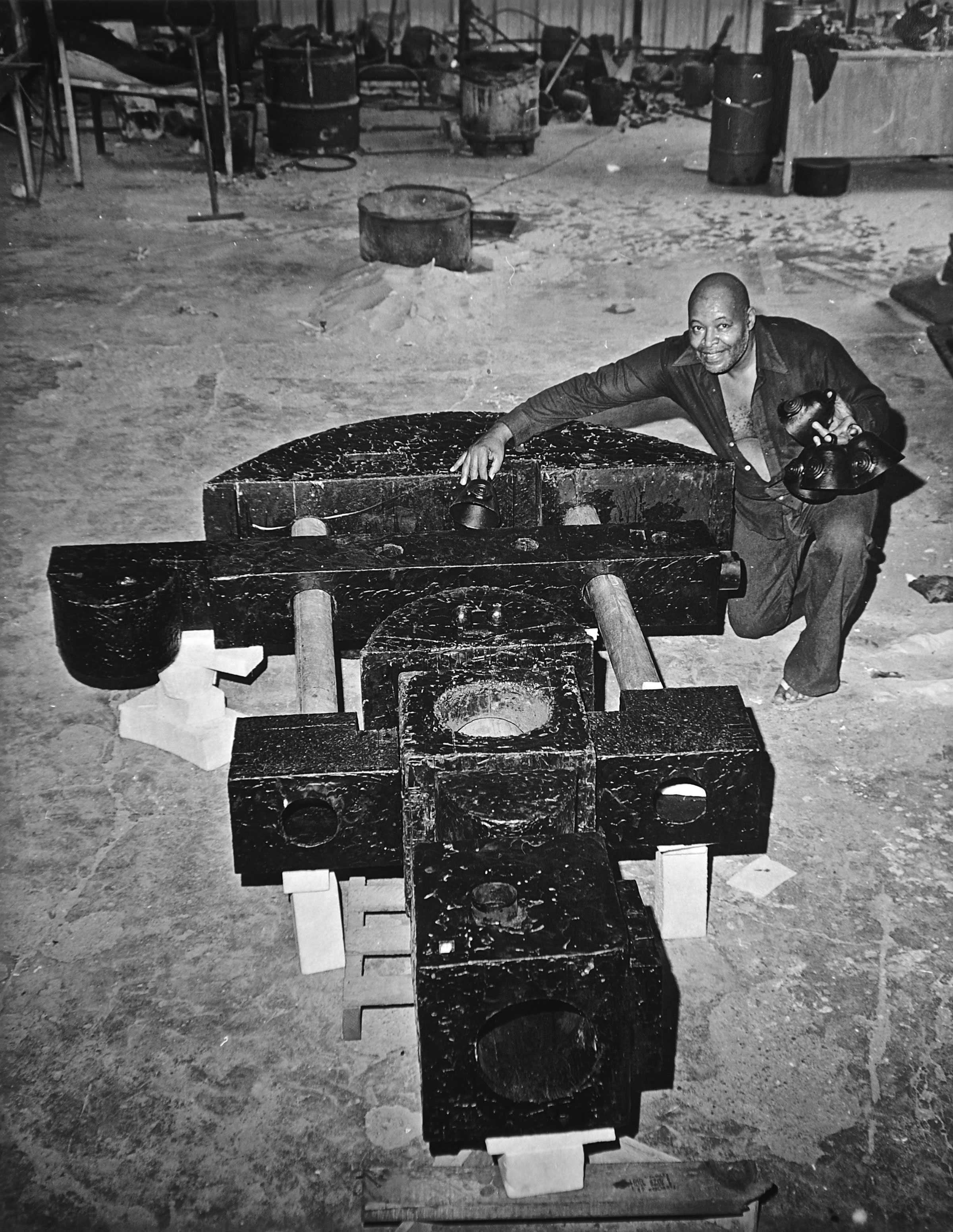 Man crouching next to a large, geometric metal sculpture in a workshop setting.