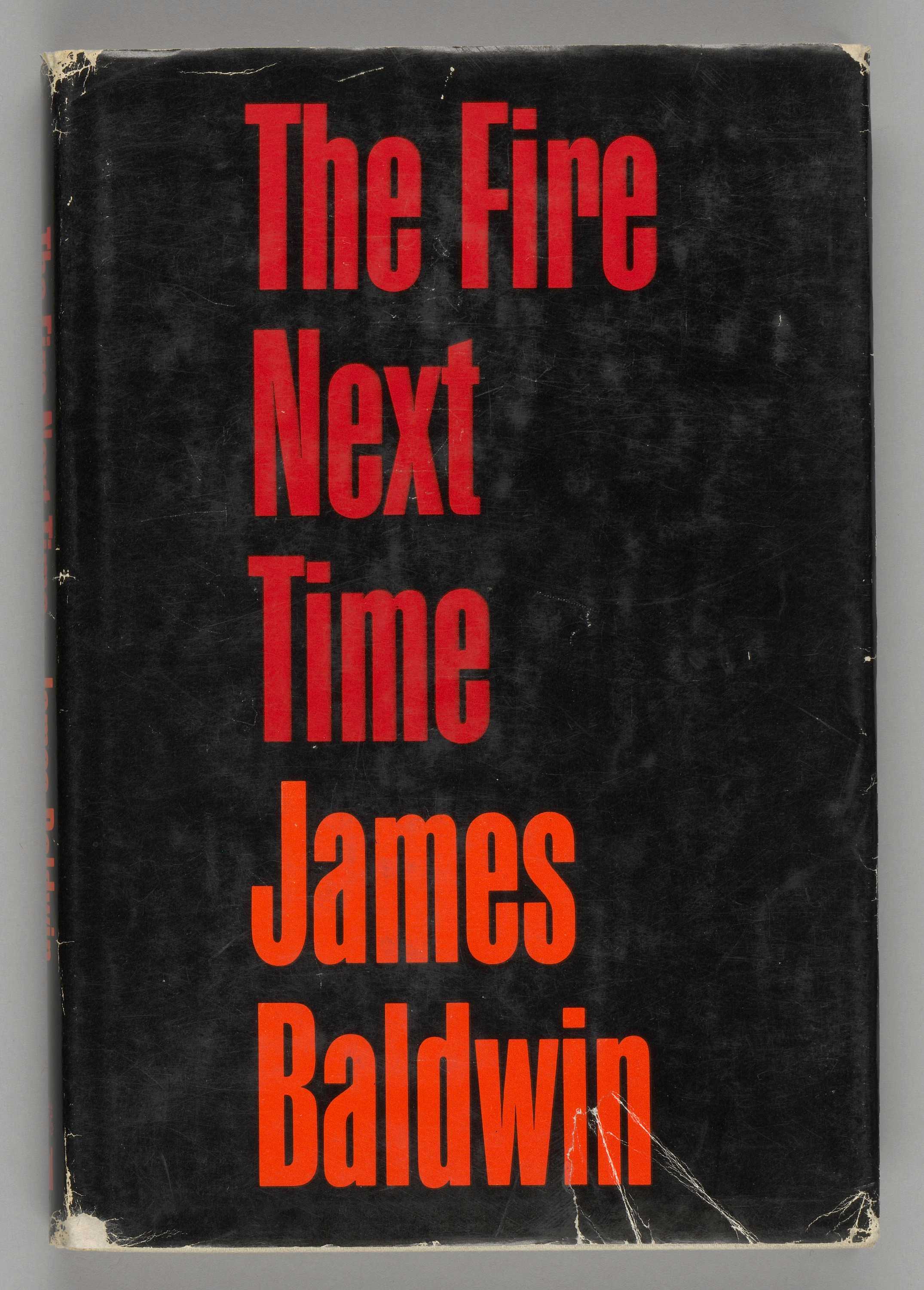 A hardback copy of The Fire Next Time by James Baldwin. The book has a paper cover with clear plastic cover over top of the book. The paper cover is black with red and orange type. The front of the paper cover reads: [The Fire / Next / Time] in red and [James / Baldwin] in orange, centered but aligned to the left. The back of the paper cover is entirely taken up by a black-and-white picture of James Baldwin. The picture is a close-up of Baldwin, who has his hands crossed in front of him and is wearing a jacket. The hardback book itself is white with red type. The interior pages, one hundred and twenty-three in total, front and back included, are off-white paper with black type, and no images. One of the interior pages has a stamp that reads: [John R. McElderry].