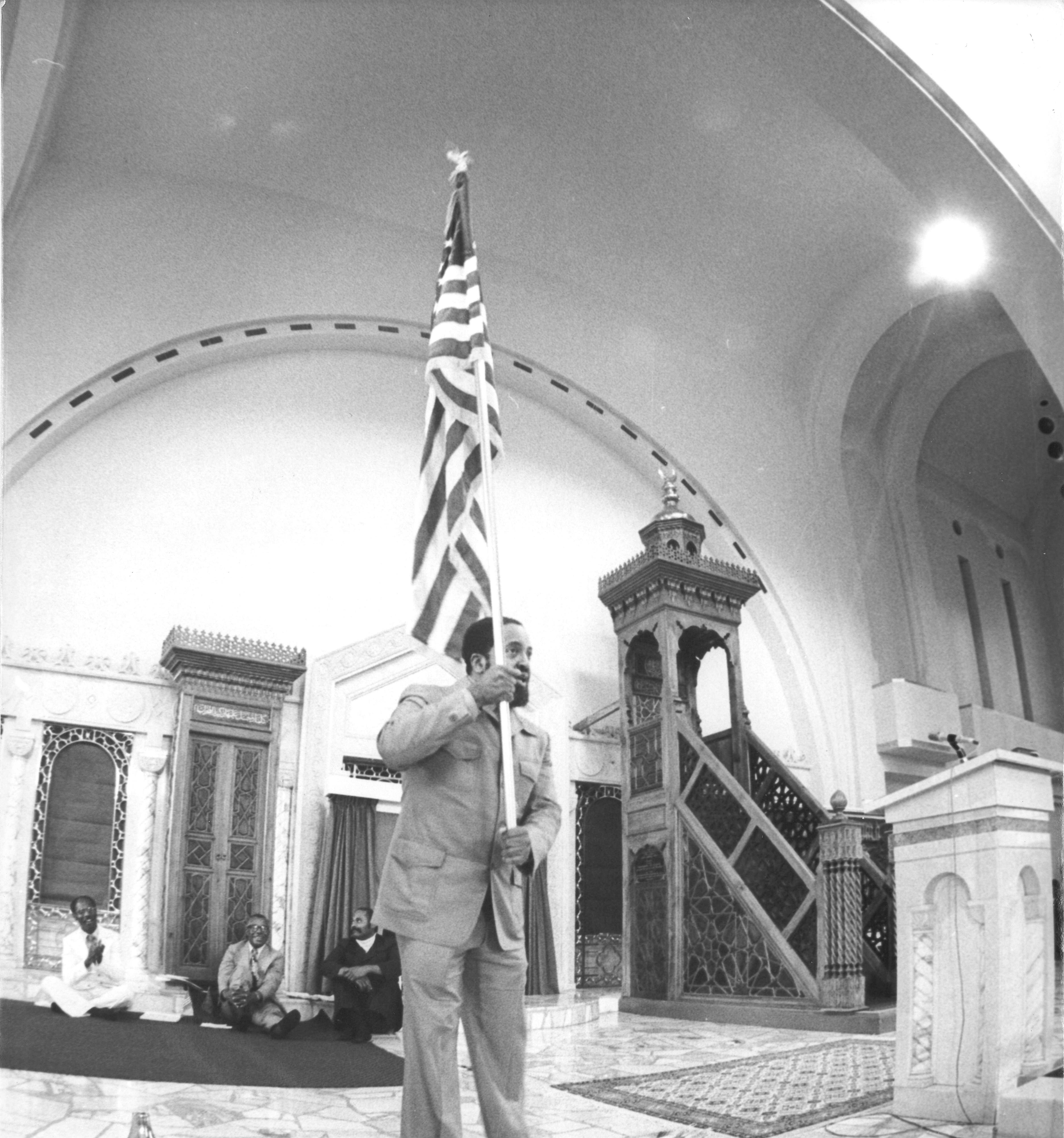 Photograph of Imam Warith Deen Mohammed carries American flag as a symbol of the nation’s obligation to promote equality for all its citizens.