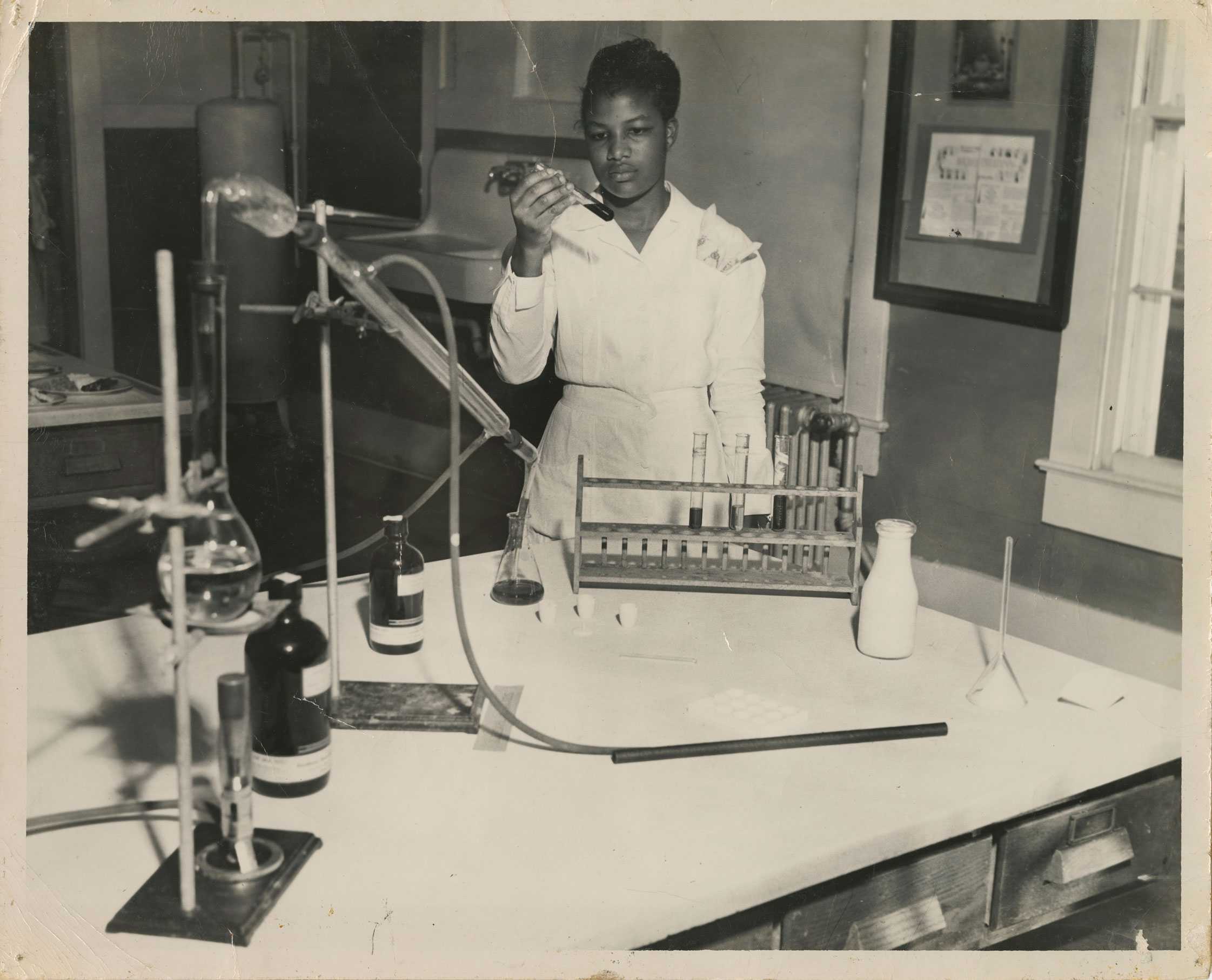 Photo of a woman in a lab coat examining a test tube in a laboratory setting with various glassware.