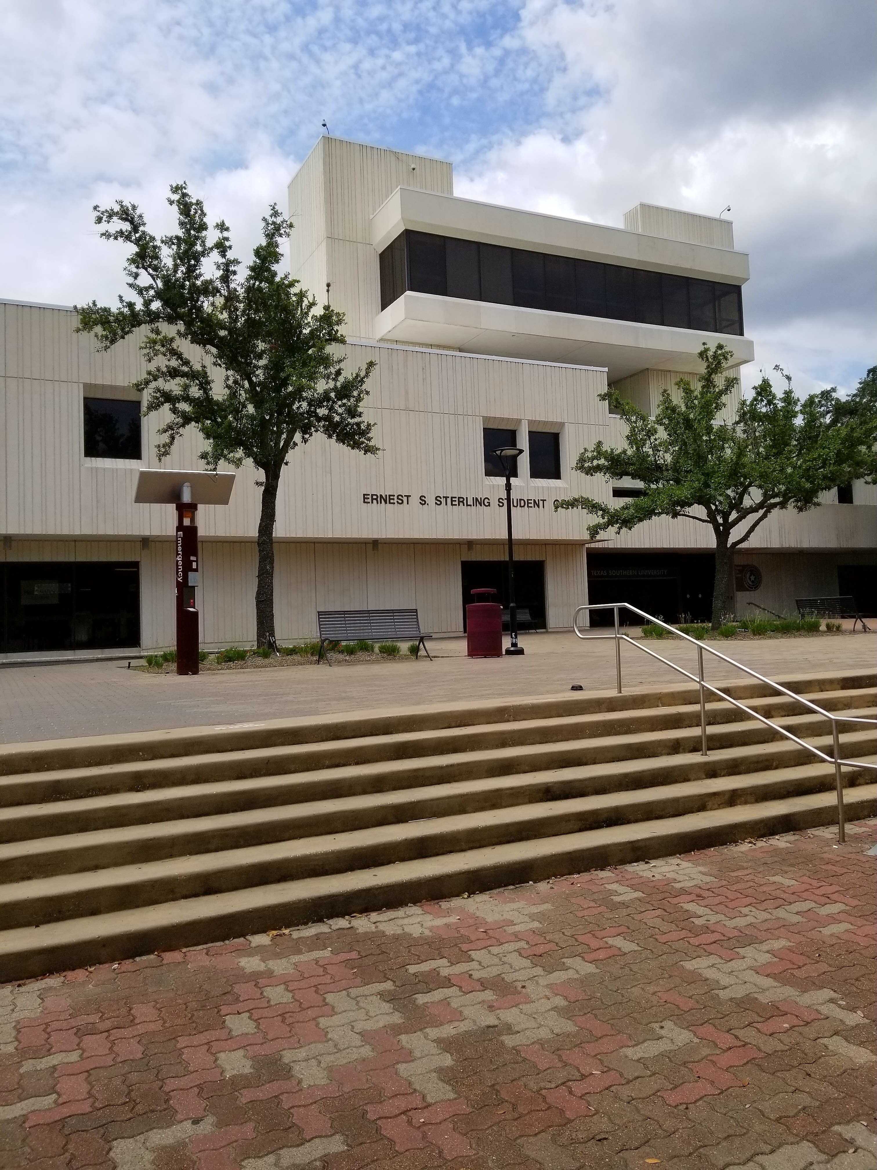 Modern courthouse building labeled 'Ernest F. Speckin Court' with white paneling, large windows, a patterned brick walkway, steps with a metal handrail, and two small trees near the entrance under a partly cloudy sky.
