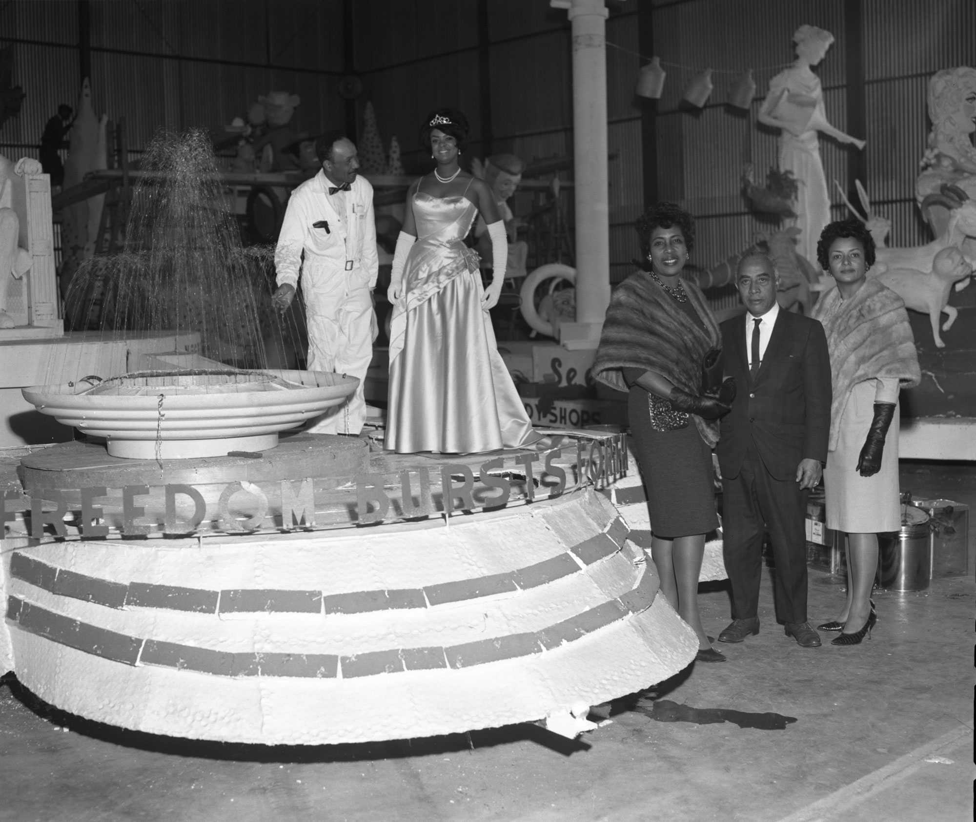 Black-and-white photo of people around a float with the text "Freedom Bursts Forth," featuring a woman in a gown and man in a work suit.