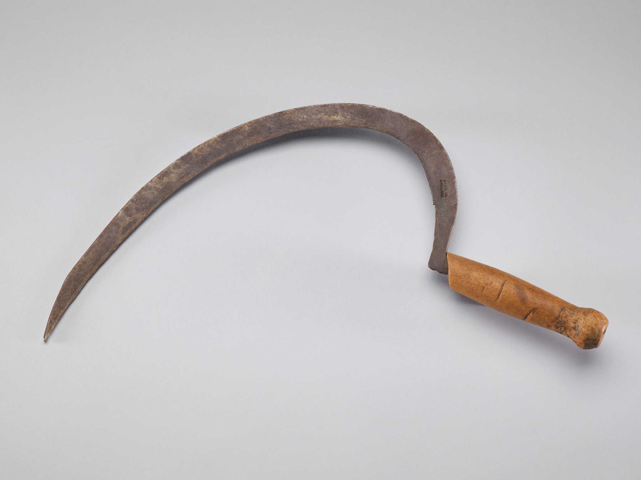 A rice sickle with a wooden handle. The sickle has a short, wooden handle with a curved grip and a rounded base. Carved on the side of the handle are the letters [W Y]. The blade is J-shaped with a serrated interior edge. Stamped on the blade near the handle is a mark that reads [MADE IN / ENGLAND]. The blade's tang protrudes from the bottom of the handle. Attached to the blade is a white paper tag with handwritten, black ink text that reads [SICKLE].