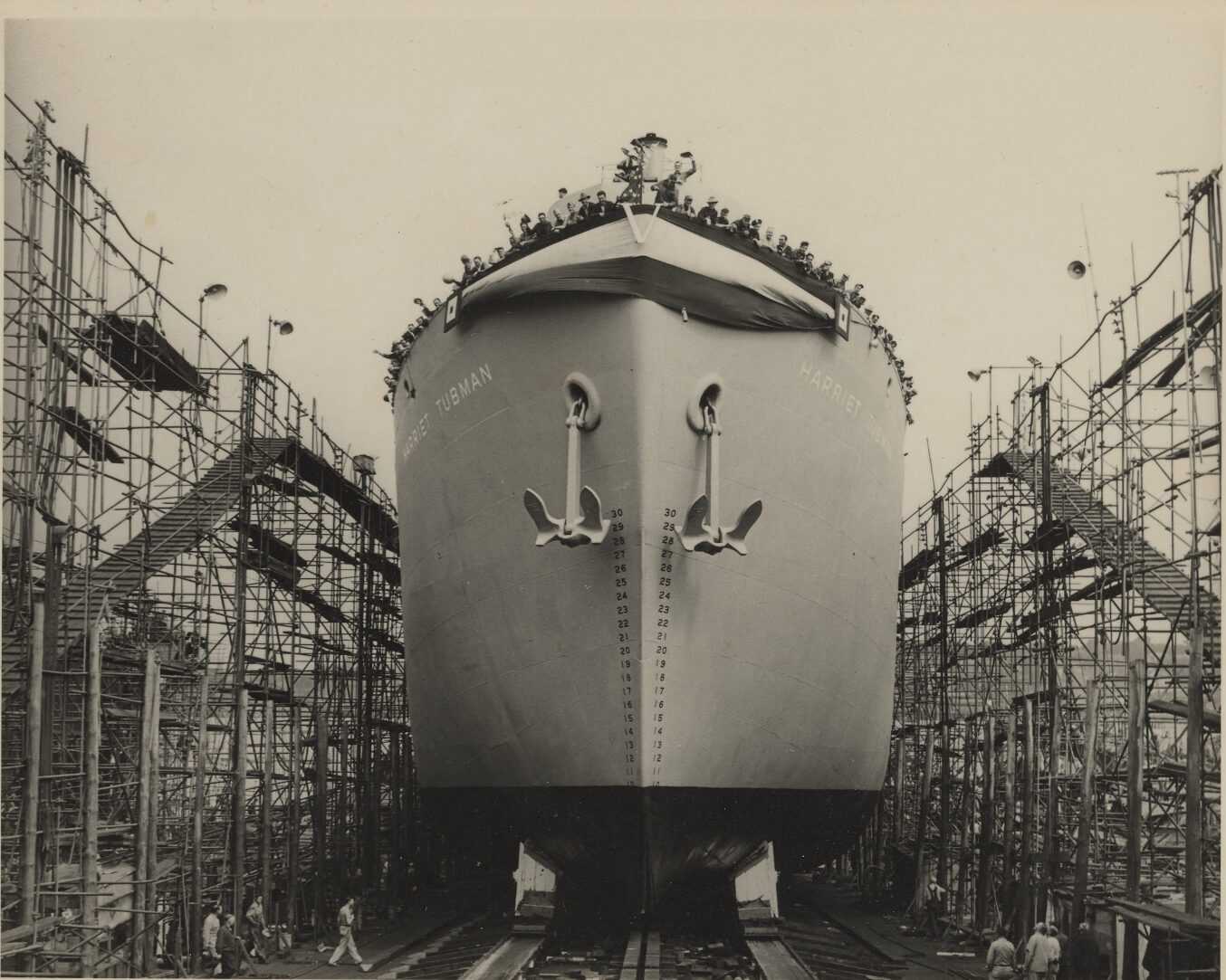 A black-and-white photograph of the liberty ship S.S. Harriet Tubman as it is launched down the slipway and into the Atlantic Ocean, mounted on a black paper page of a bound photograph album commemorating the event owned by Harriet Tubman's niece, Eva Stewart Northrup. The bow of the ship is depicted in the center of the image, surrounded by scaffolding. Two raised anchors feature prominently on either side of the ship and at the top is a striped banner with a large "V" victory symbol at the center. On each side of the ship are large letters reading [HARRIET TUBMAN]. The deck of the ship is loaded with sailors, shipwrights and others, many waving their hats in the air. There are also several men standing on either side of the shipway. There are no marks or inscriptions, front or back.